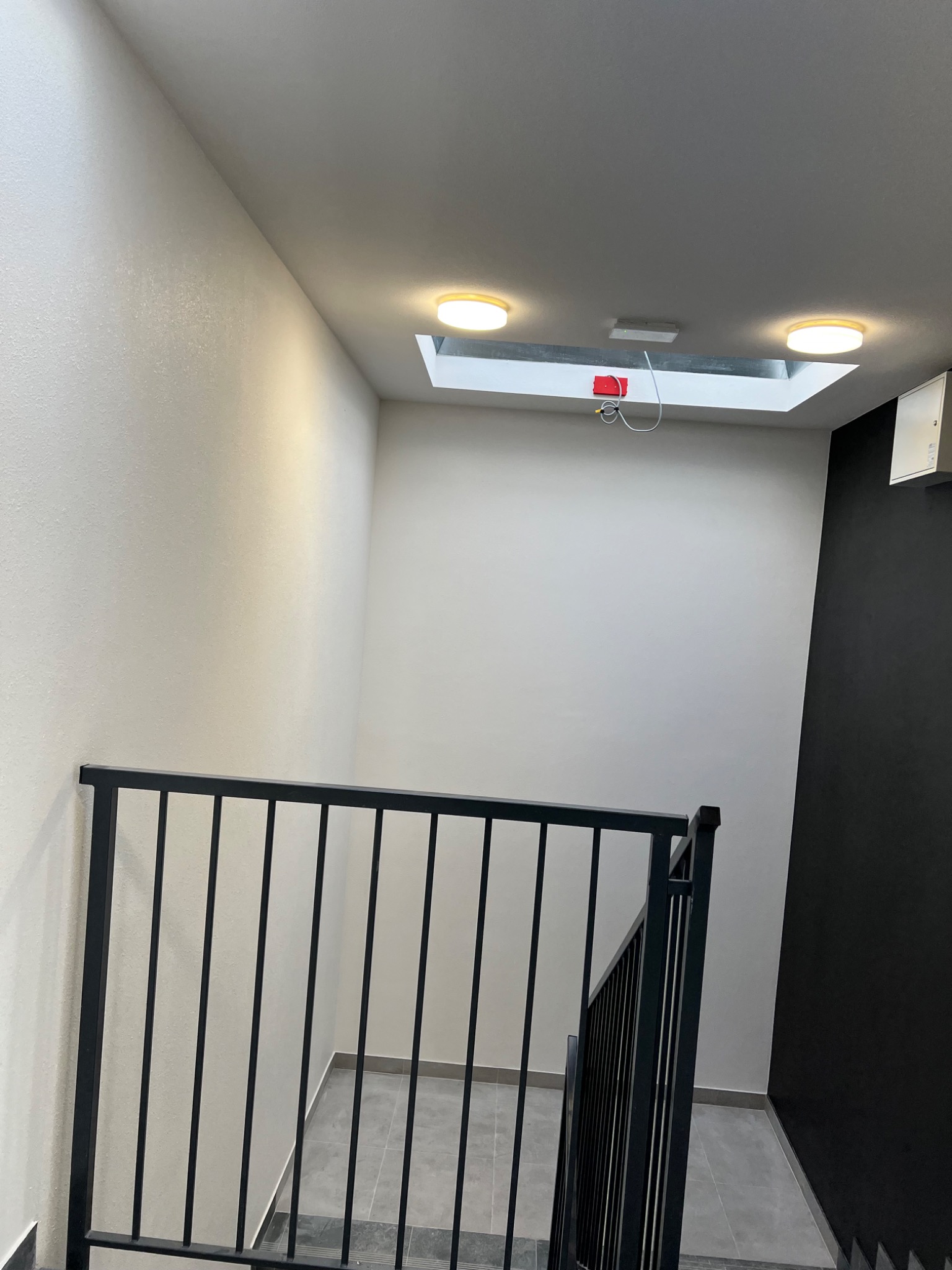 Klatka schodowa z czarną metalową balustradą, białe ściany i sufit z wbudowanym oświetleniem, fragment czarnej ściany po prawej stronie, szare płytki podłogowe.