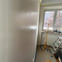 Firma Remontowo-Budowlana DAW-BUD Dawid Bala - Świeżo pomalowana, mokra ściana w pokoju z oknem, drabina, kuweta z farbą i lampa budowlana na trójnogu.