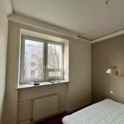 Firma Remontowo-Budowlana DAW-BUD Dawid Bala - Jasne wnętrze pokoju po malowaniu, widoczne świeżo pomalowane ściany w odcieniach beżu i bieli, biała rama okienna z widokiem na budynek i drzewa, grzejnik pod oknem, fragment łóżka z białą...