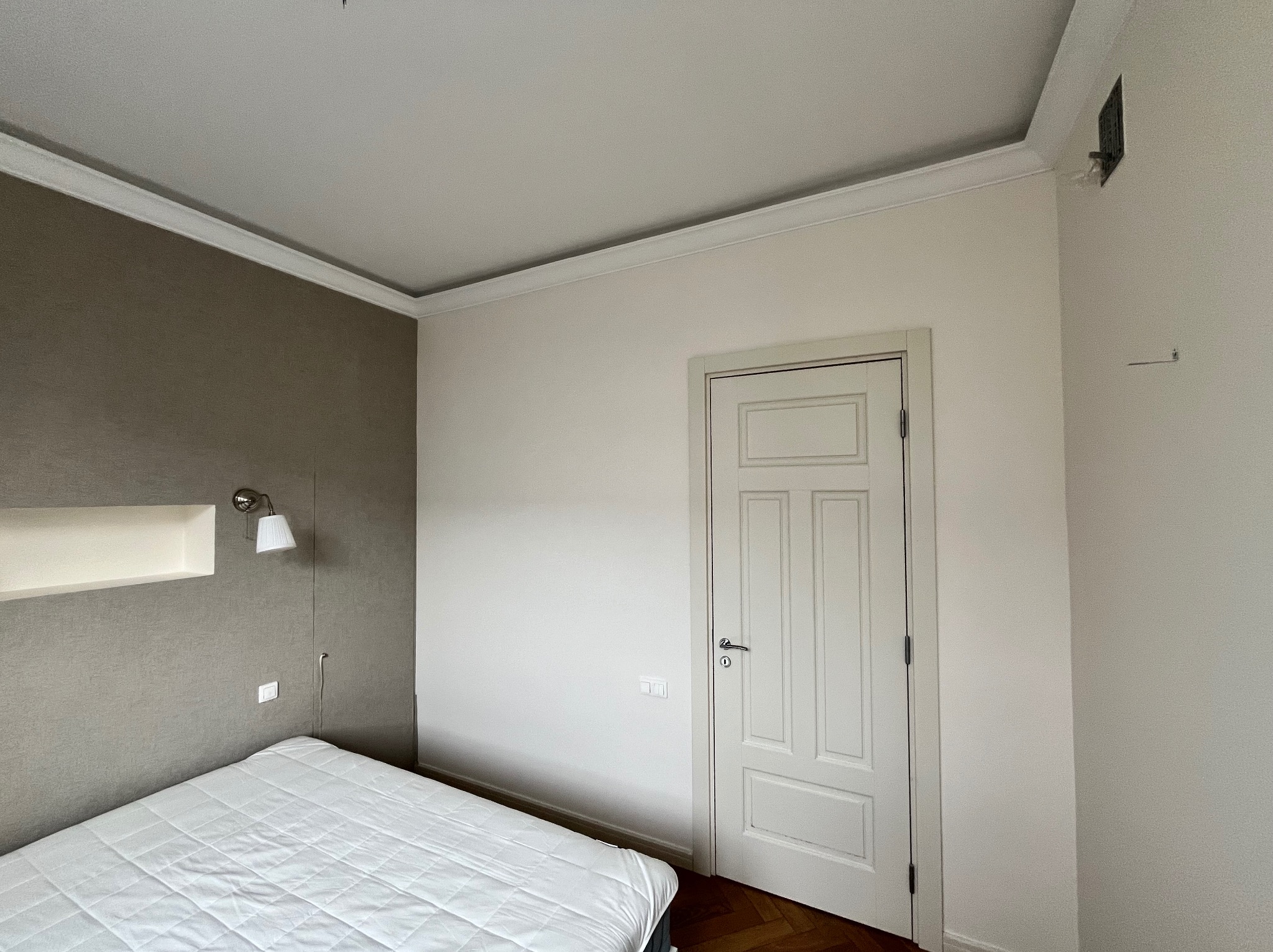 Ściana w sypialni pomalowana na dwa kolory: szary z lewej strony i biały z prawej, z białą, klasyczną stolarką drzwiową i parkietem w jodełkę.