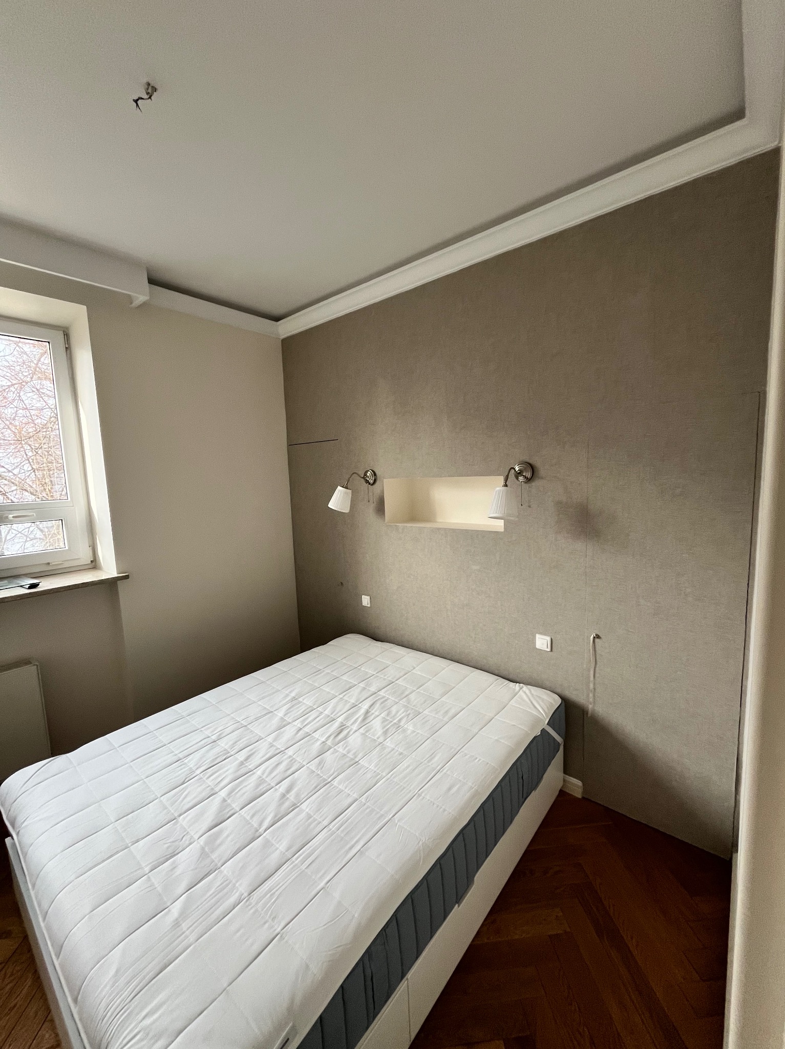 Pokój przed remontem: Łóżko z białym materacem na tle ściany w kolorze taupe, z widocznymi śladami po zdjętych elementach, lampkami nocnymi i niezamaskowanymi przewodami.