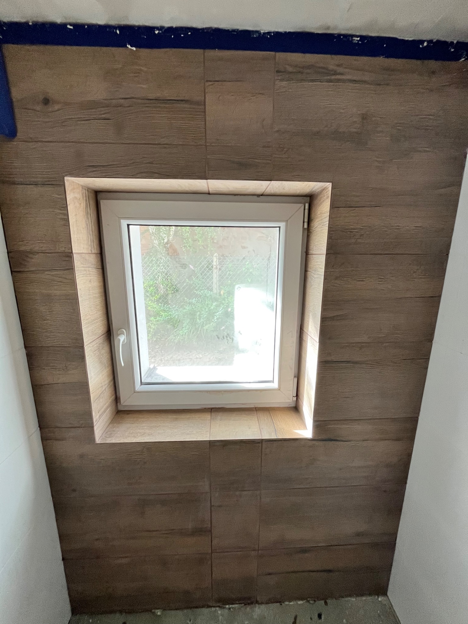 Okno w obudowie z płytek imitujących drewno, widok z dołu. Płytki ułożone na skosie poddasza, białe ściany po bokach, niebieska taśma malarska przy suficie.