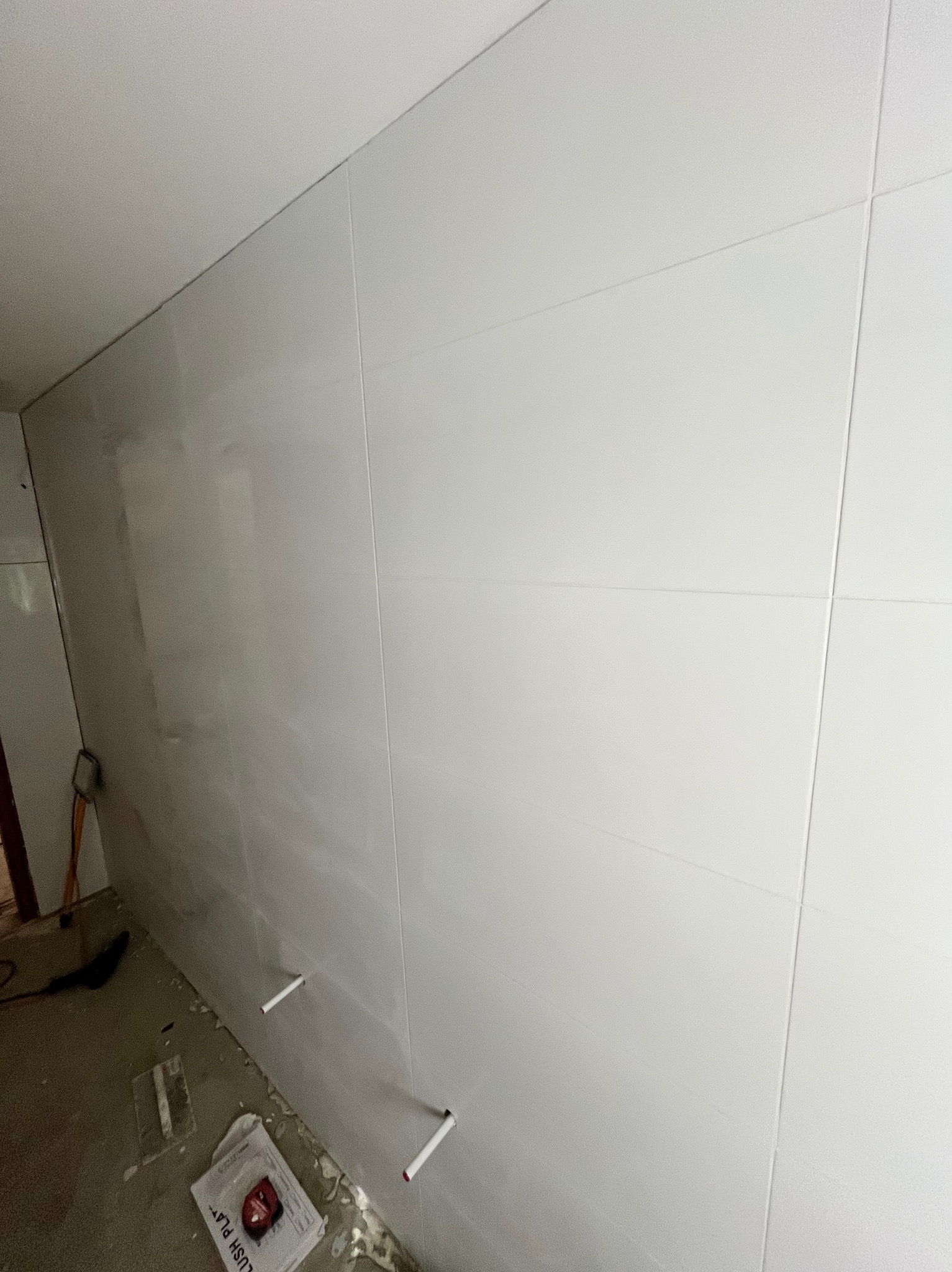 Ściana wyłożona białymi, błyszczącymi płytkami ceramicznymi, z widocznymi rurami instalacji. Na podłodze narzędzia glazurnicze i resztki materiałów.