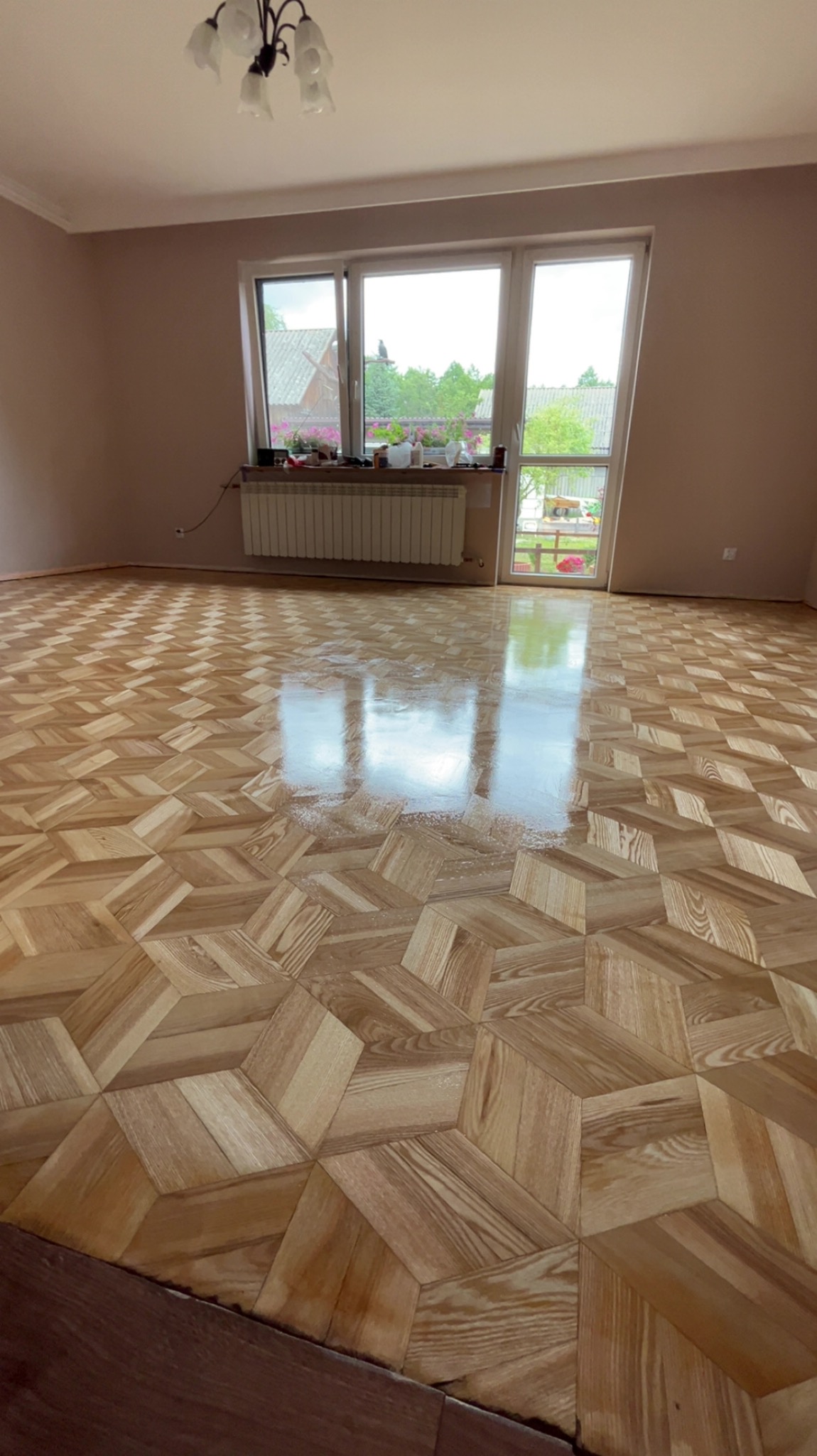 Błyszcząca, drewniana podłoga parkietowa w pustym pokoju z oknem i grzejnikiem. Widoczne refleksy światła na lakierowanej powierzchni. Jasne ściany i sufit.