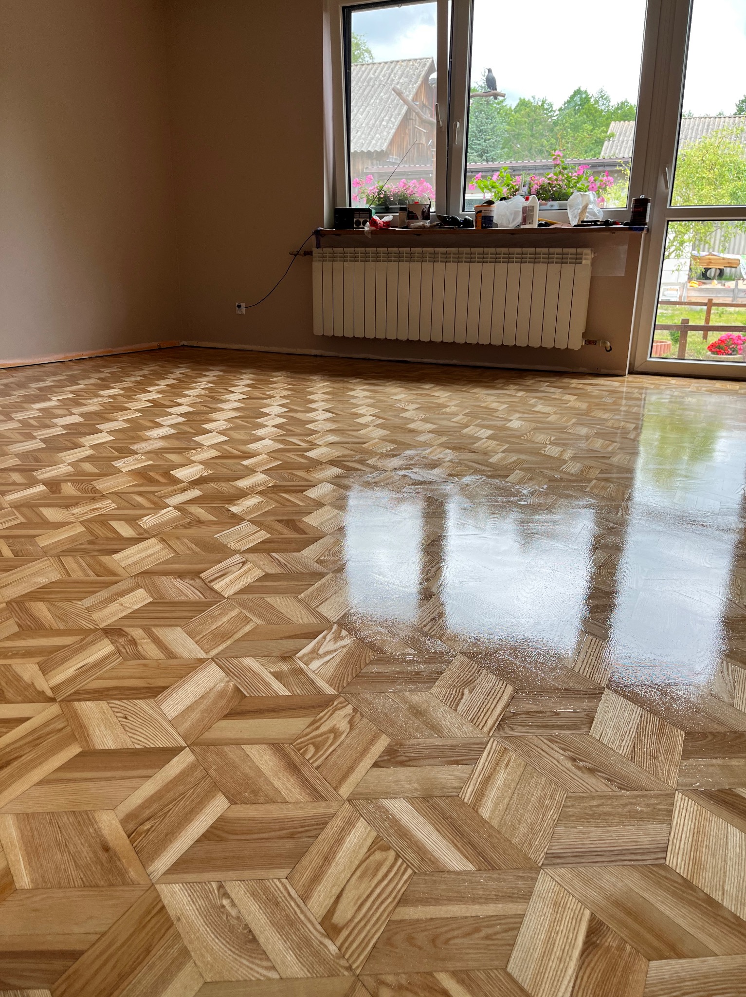 Parkiet ułożony we wzór 3D, świeżo polakierowany, odbija światło. Widoczne okno z kwiatami i grzejnik. Ściany w neutralnym beżu. Listwa przypodłogowa w kolorze podłogi.