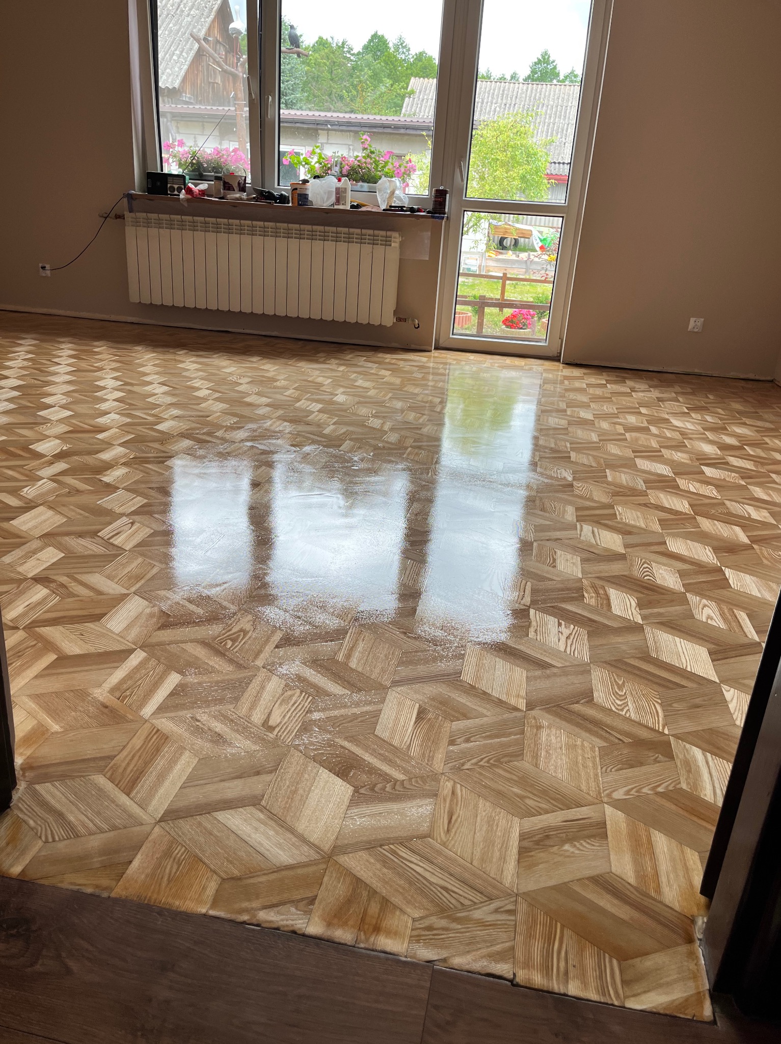 Parkiet ułożony w geometryczny wzór, świeżo po lakierowaniu, z widocznymi refleksami światła. W tle okno z widokiem na zieleń i fragment grzejnika.