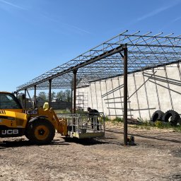 WELD SERVICE USŁUGI SPAWALNICZE - Montaż stalowej konstrukcji hali z użyciem podnośnika koszowego JCB, widoczne elementy konstrukcyjne i pracownik w koszu na tle jasnego nieba.