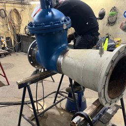 WELD SERVICE USŁUGI SPAWALNICZE - Fragment rurociągu z zaworem, spawany przez pracownika w kombinezonie, w warsztacie z narzędziami i akcesoriami spawalniczymi.