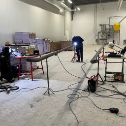 WELD SERVICE USŁUGI SPAWALNICZE - Prace spawalnicze w hali produkcyjnej: spawanie rur na statywach, widoczne iskry i sprzęt spawalniczy, w tle zaplecze magazynowe.