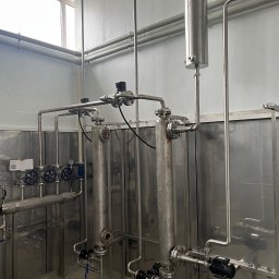 WELD SERVICE USŁUGI SPAWALNICZE - Instalacja przemysłowa z rurami ze stali nierdzewnej, zaworami i zbiornikami w pomieszczeniu z oknem. Widoczne złącza kołnierzowe i elementy mocujące.