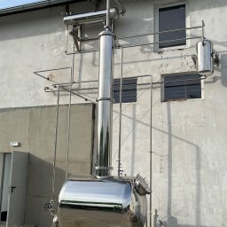 WELD SERVICE USŁUGI SPAWALNICZE - Instalacja przemysłowa ze stali nierdzewnej, zbiornik ciśnieniowy z rurami na tle budynku z oknami. Błyszcząca powierzchnia metalu odbija otoczenie.