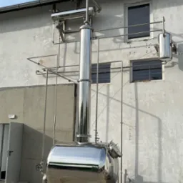 Instalacja przemysłowa ze stali nierdzewnej, zbiornik ciśnieniowy z rurami na tle budynku z oknami. Błyszcząca powierzchnia metalu odbija otoczenie.