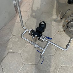 WELD SERVICE USŁUGI SPAWALNICZE - Metalowa instalacja z zaworami i rurami, widok z góry. Połączenia gwintowane, element zbiornika. Posadzka z szarej kostki brukowej w kształcie sześciokątów.