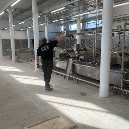 WELD SERVICE USŁUGI SPAWALNICZE - Pracownik w firmowej odzieży, kontroluje metalową instalację przemysłową w przestronnej hali z białymi kolumnami i oświetleniem. Widoczny fragment podłogi.