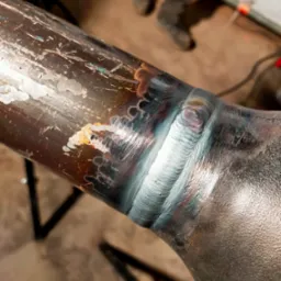 Spoina na rurze metalowej, widoczne ślady po obróbce i przypalenia, w tle zarys sylwetki osoby i elementy warsztatu.