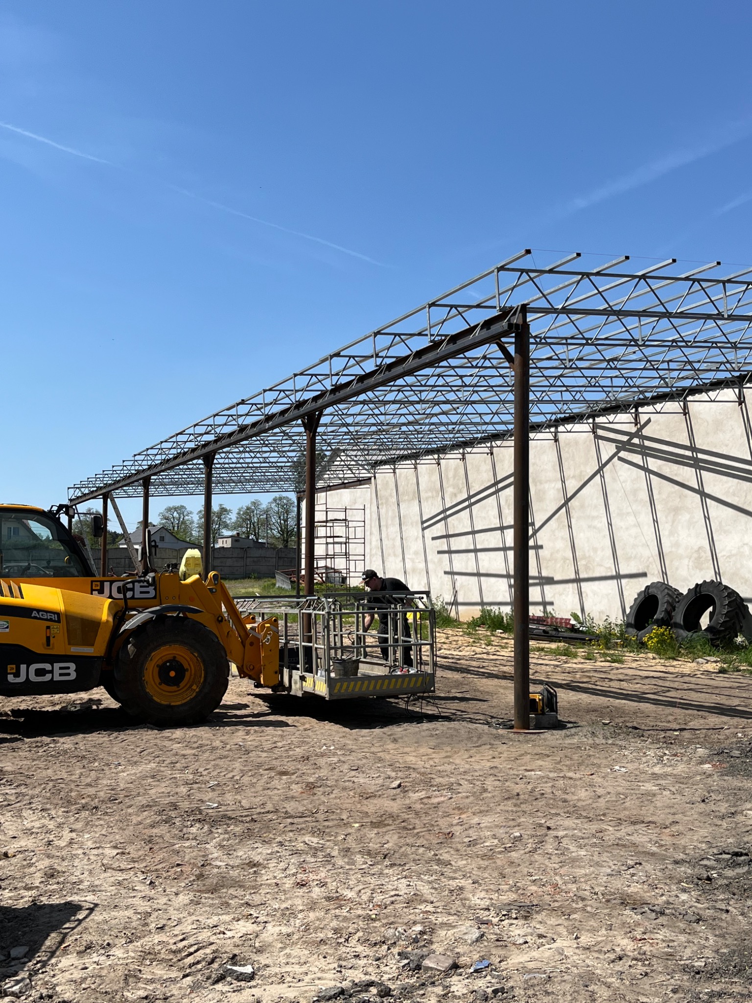 Montaż stalowej konstrukcji hali z użyciem podnośnika koszowego JCB, widoczne elementy konstrukcyjne i pracownik w koszu na tle jasnego nieba.