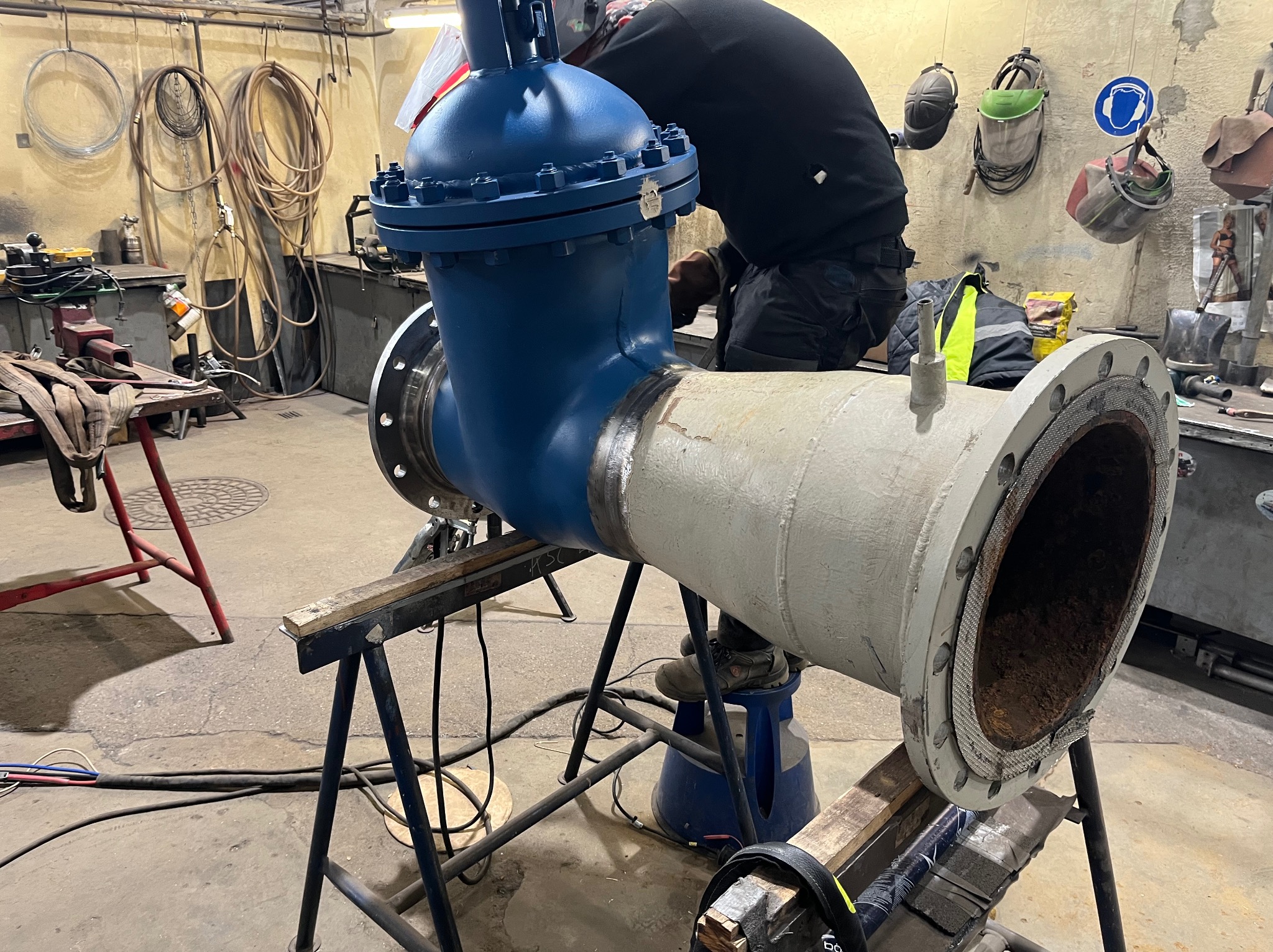 Fragment rurociągu z zaworem, spawany przez pracownika w kombinezonie, w warsztacie z narzędziami i akcesoriami spawalniczymi.