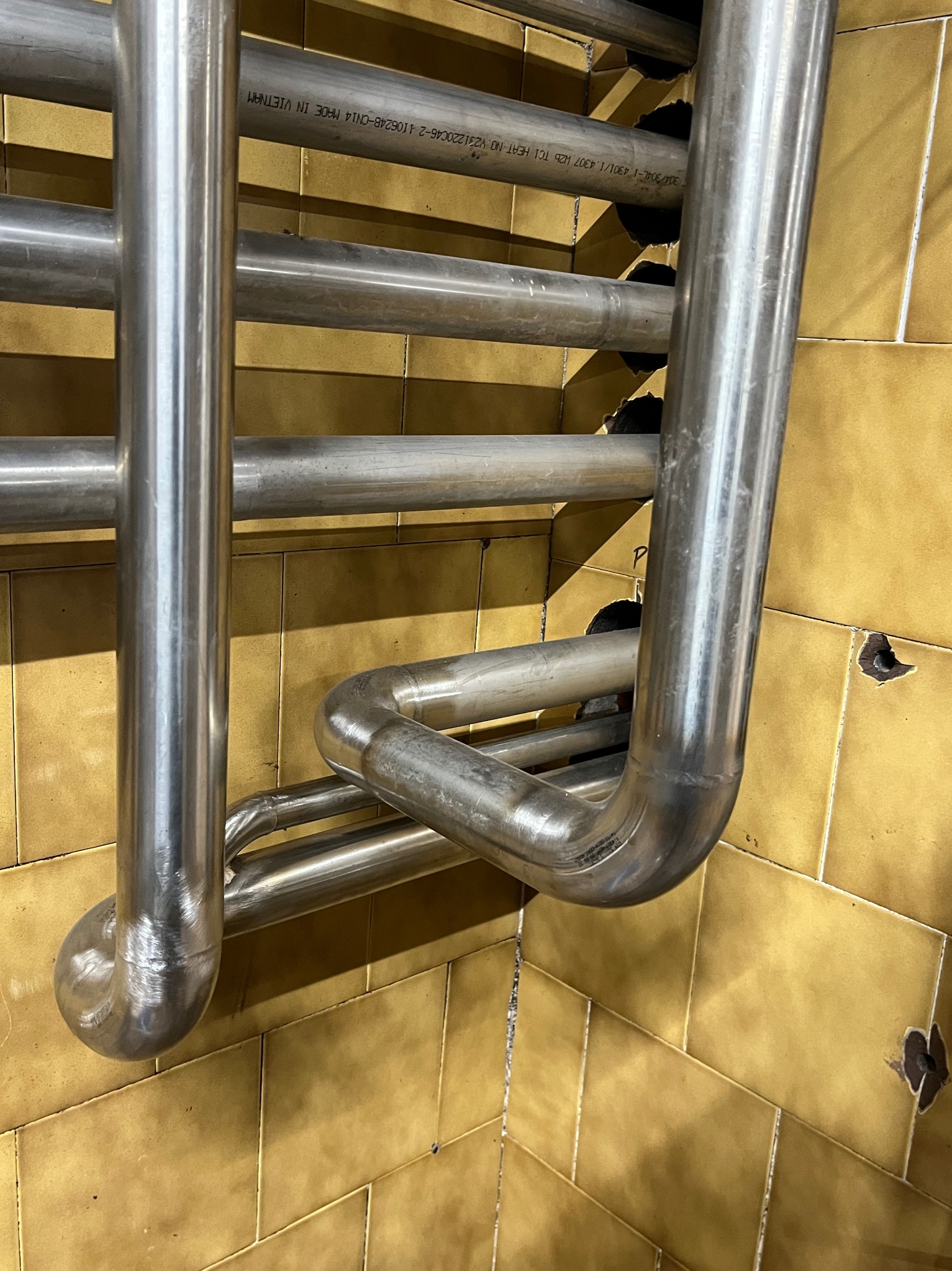 Fragment konstrukcji ze stalowych rur, prawdopodobnie grzejnika łazienkowego, zamontowanego na ścianie wyłożonej żółtymi, błyszczącymi płytkami. Widoczne ślady po wierceniu w fugach.