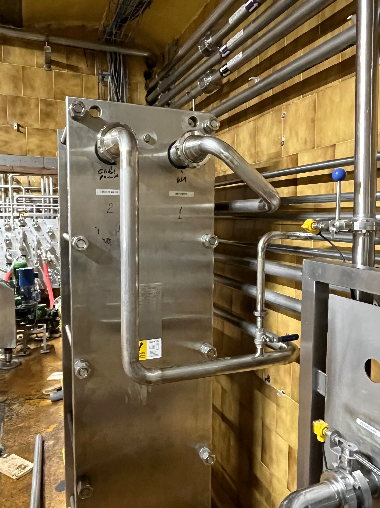 Metalowa instalacja przemysłowa z rurami i zaworami, widoczne napisy i oznaczenia na elementach, tło z płytek ceramicznych.