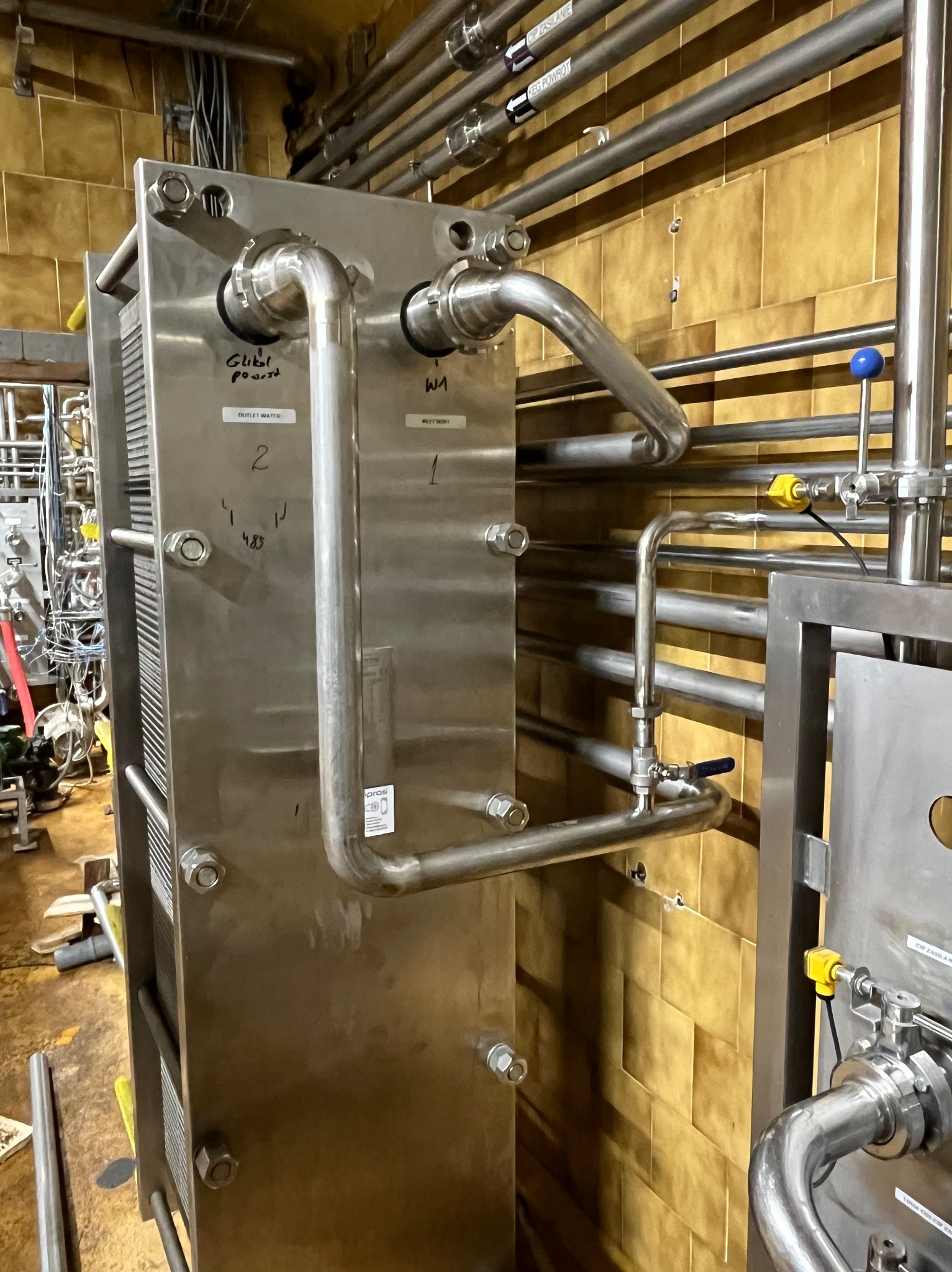 Płytowy wymiennik ciepła ze stali nierdzewnej z rurami, widoczne przyłącza, zawory i napisy informacyjne na tle wyłożonej kafelkami ściany.