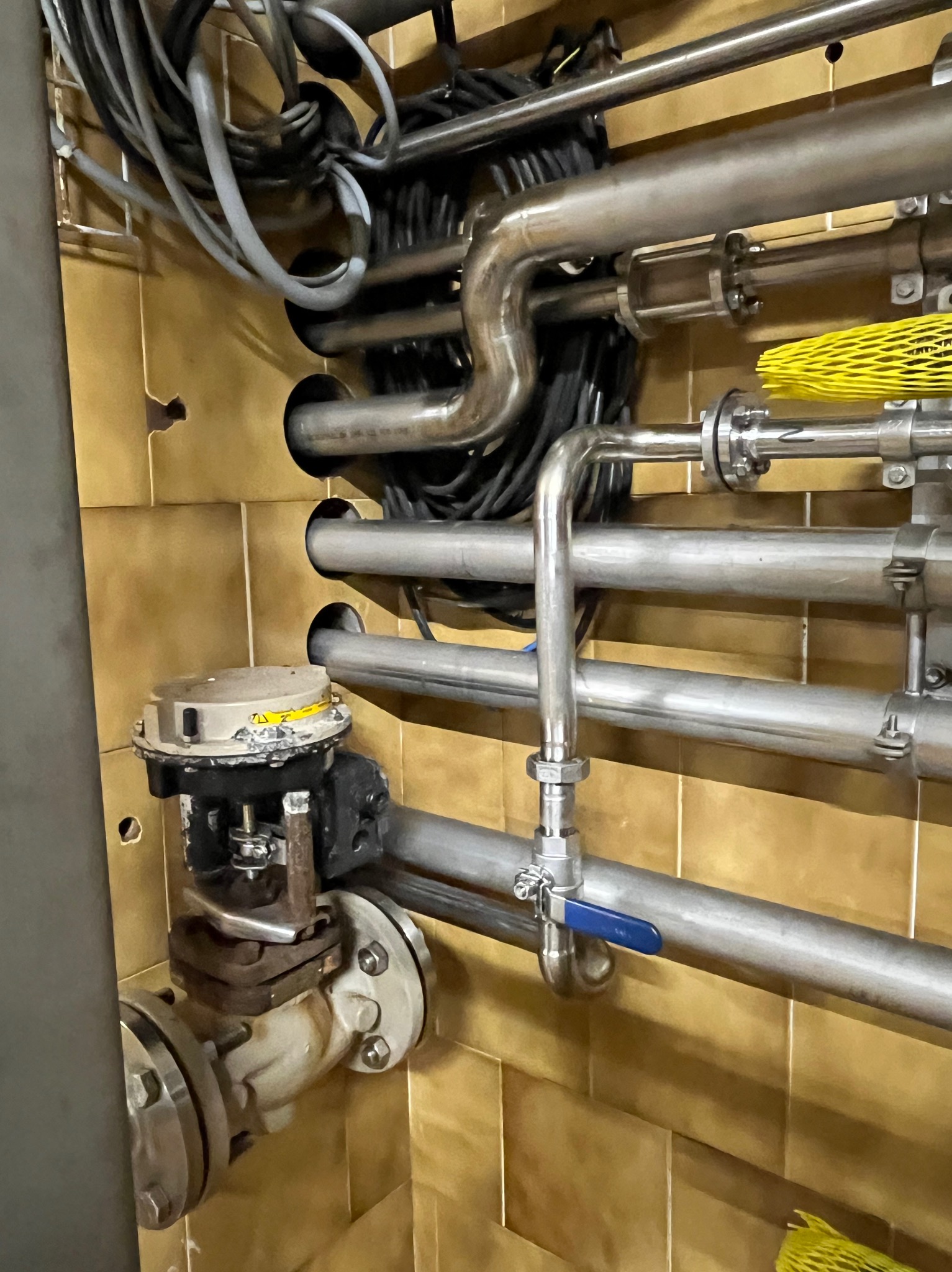 Złożony system rur ze stali nierdzewnej z zaworami i przewodami elektrycznymi na tle kafelkowej ściany.