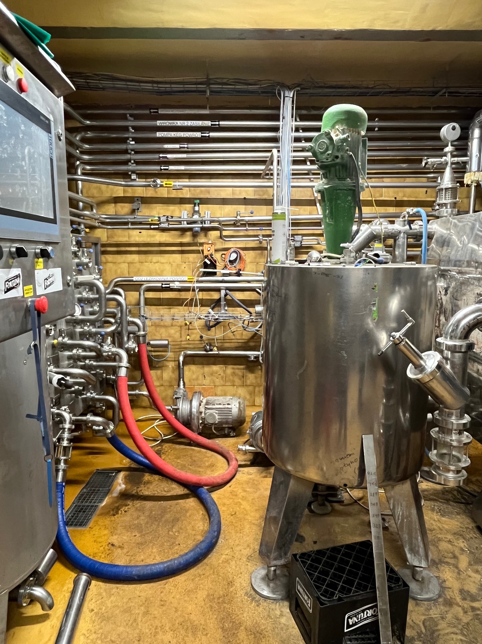 Wnętrze przemysłowe z instalacją ze stali nierdzewnej, zbiornikiem z zielonym silnikiem na górze, rurami i przewodami, oraz panelem sterowania z etykietami 'Fortuna' i napisami w języku polskim...