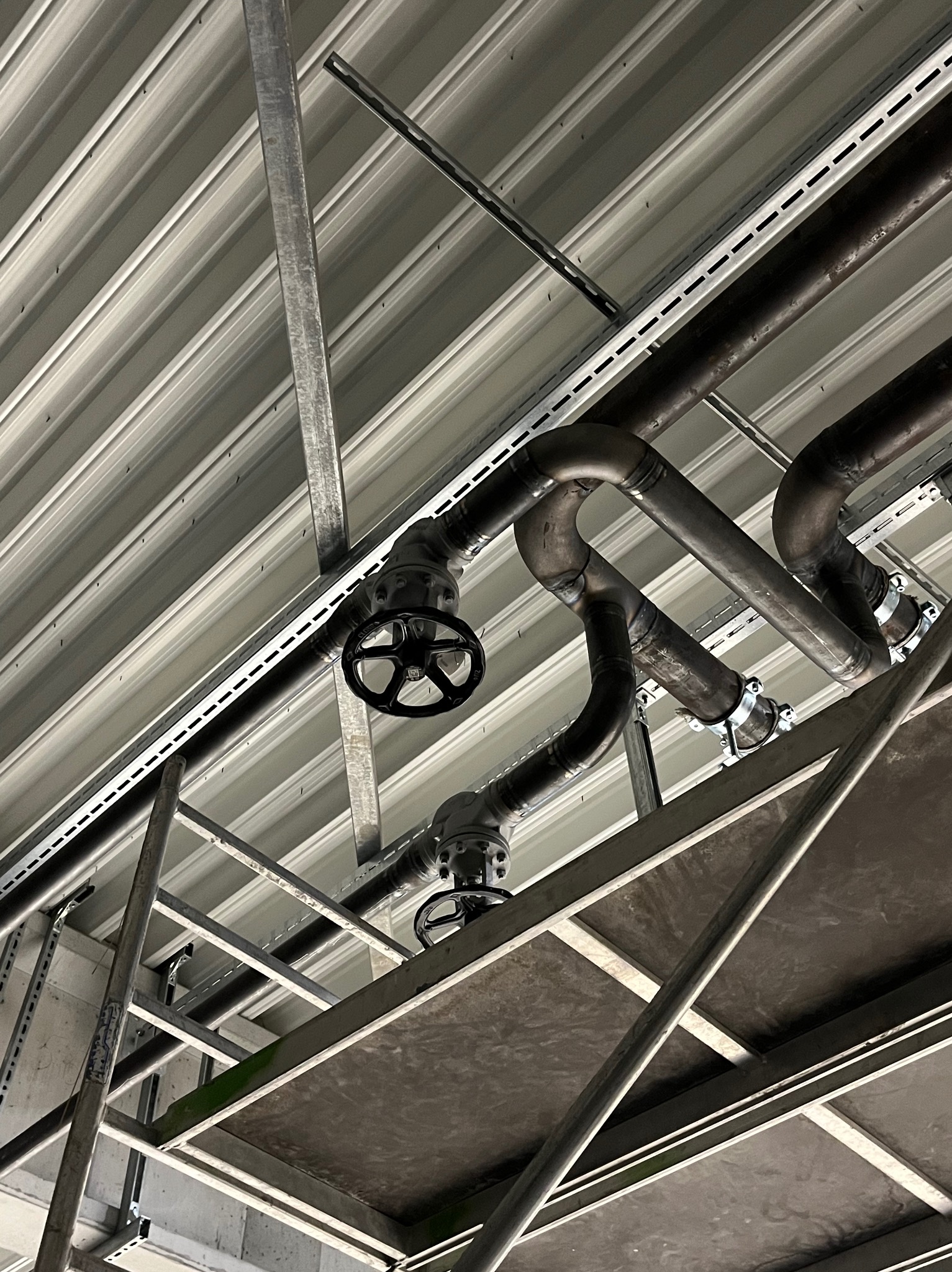 Widok z dołu na rurociąg przemysłowy z zaworami, zamocowany pod stropem hali, widoczne łączenia spawane i obejmy mocujące.