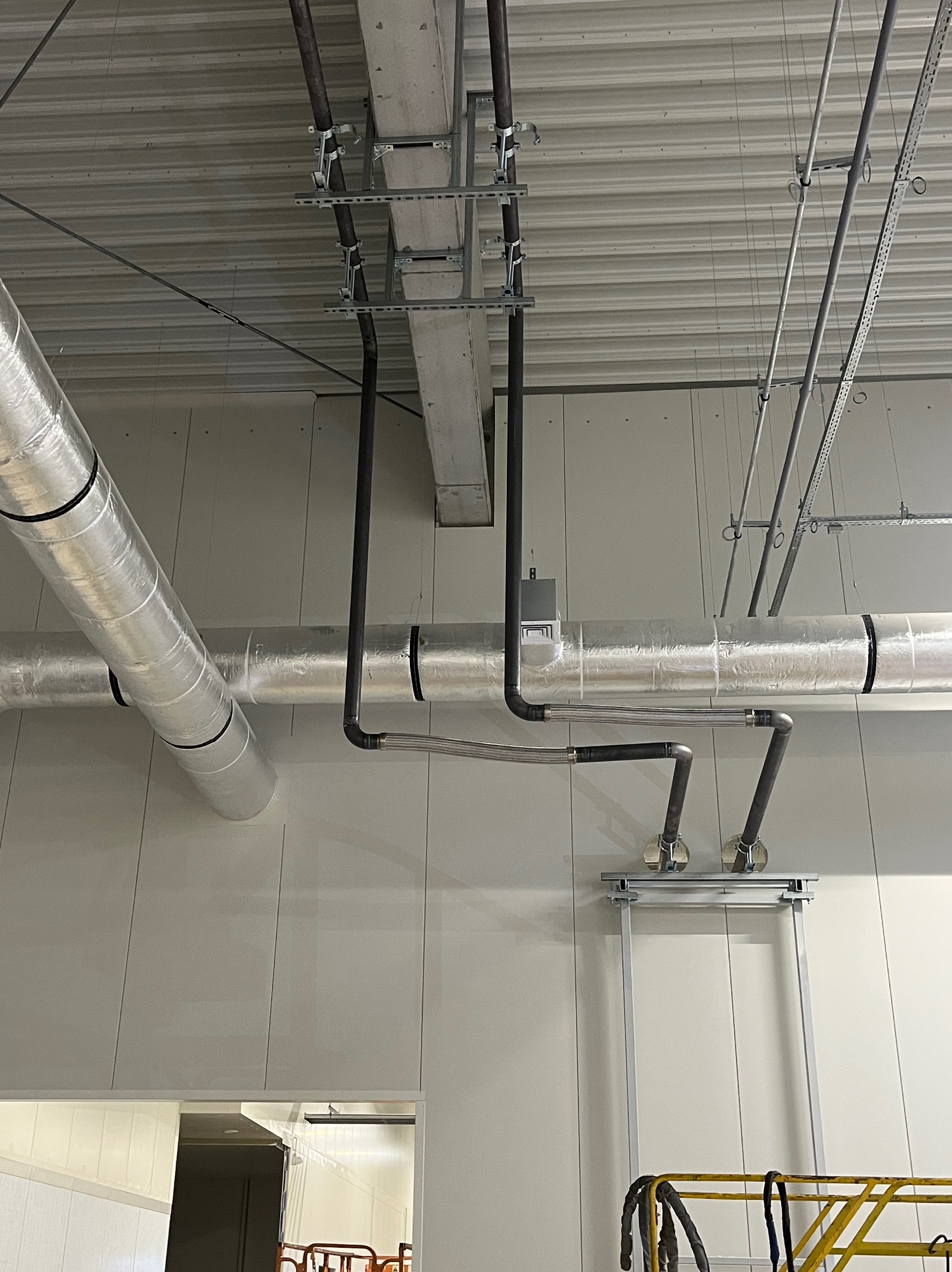 Instalacja przemysłowa: srebrna izolacja termiczna rur, czarne rury, metalowe wsporniki pod sufitem, fragment podnośnika w rogu kadru.