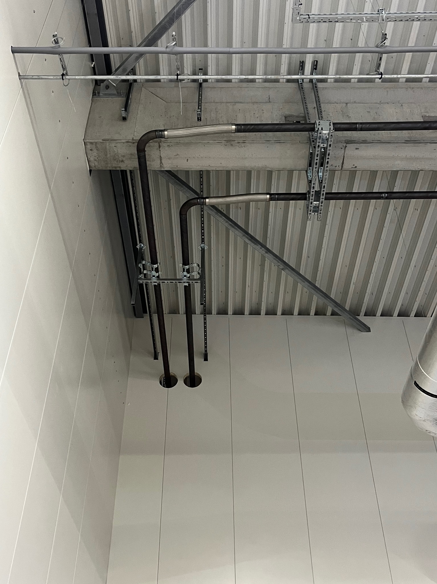 Wnętrze hali w trakcie budowy: widoczne rury instalacyjne, metalowe konstrukcje podtrzymujące pod sufitem, oraz fragment ściany z paneli.