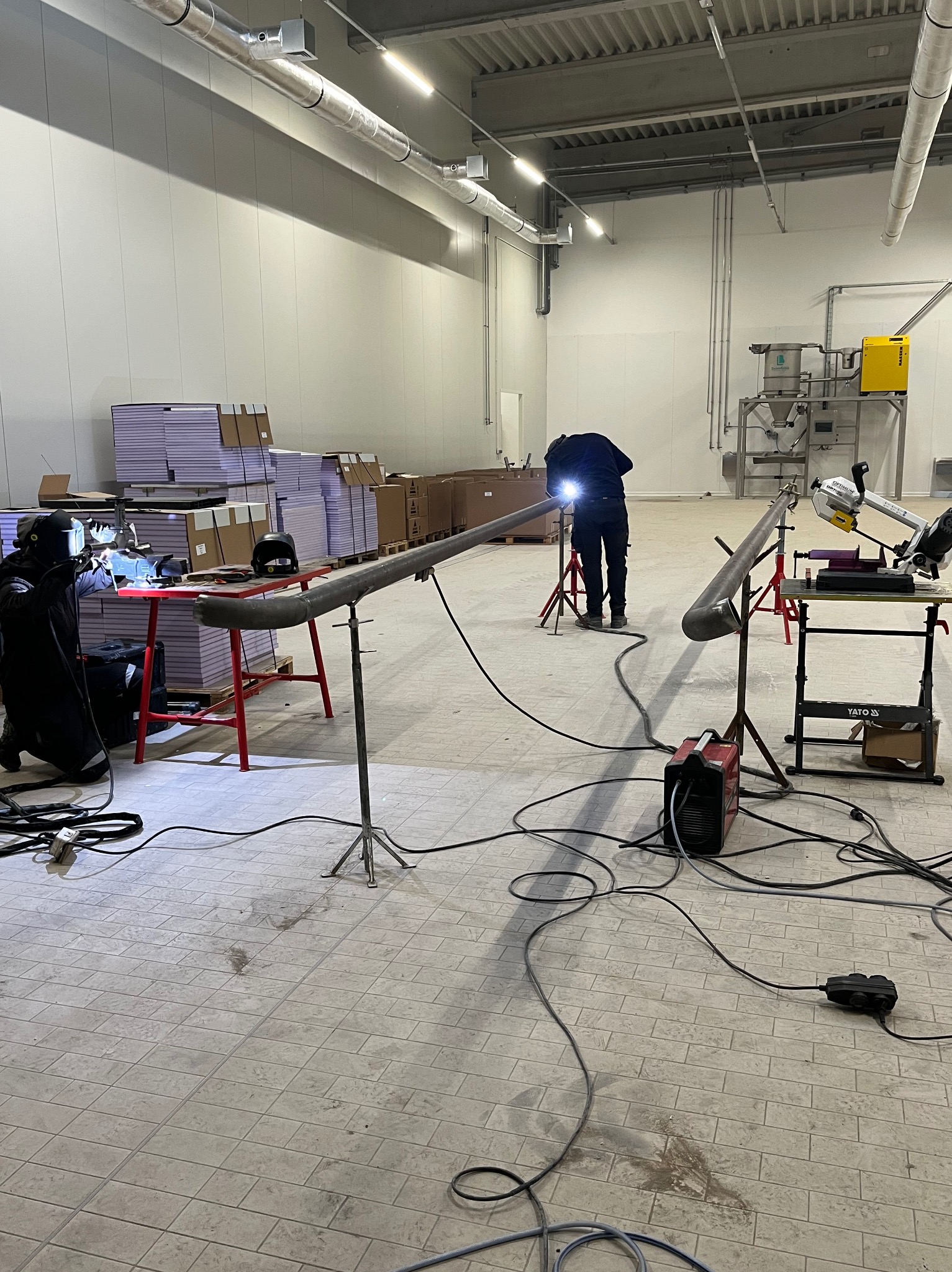 Prace spawalnicze w hali produkcyjnej: spawanie rur na statywach, widoczne iskry i sprzęt spawalniczy, w tle zaplecze magazynowe.