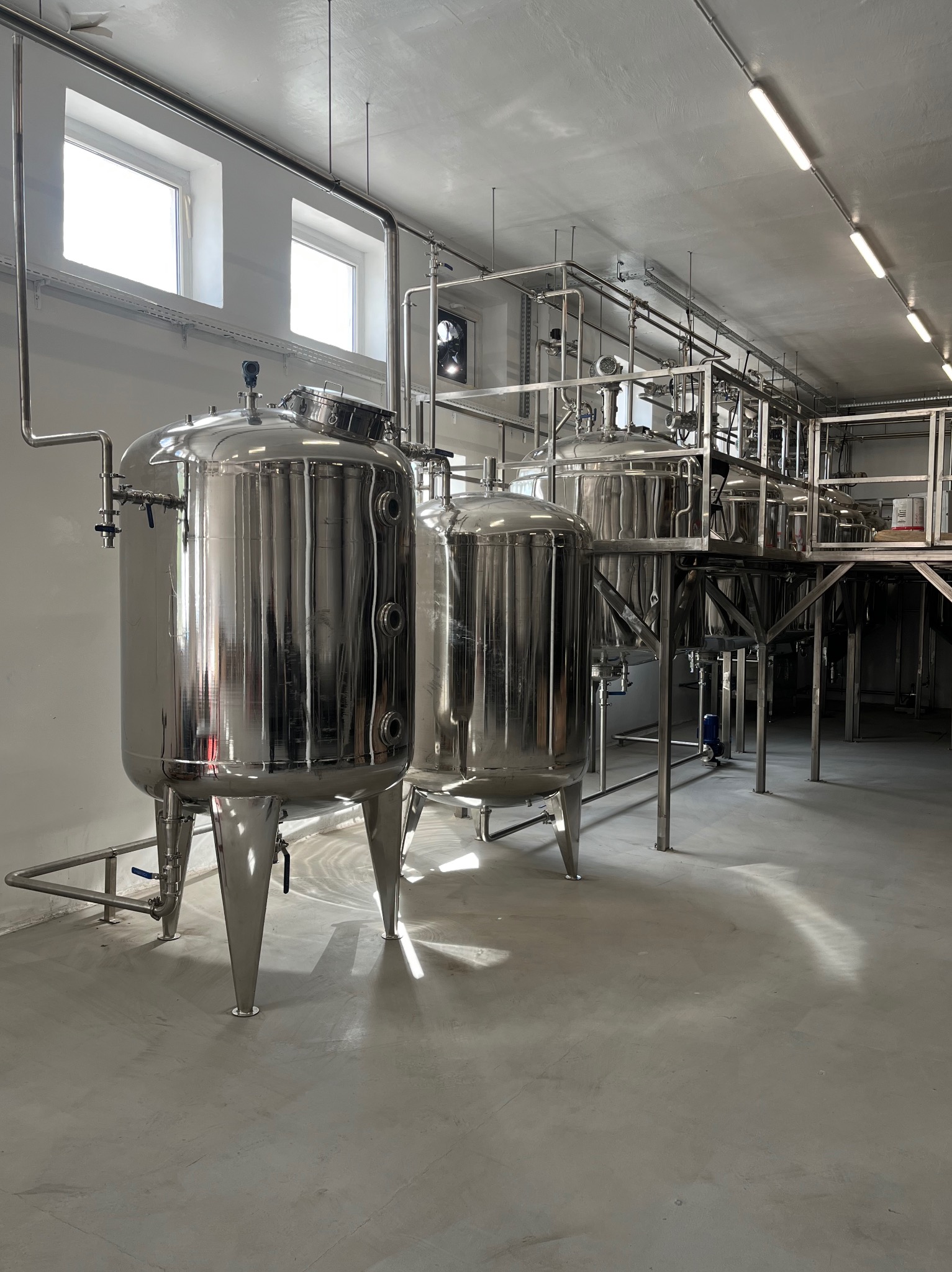 Błyszczące, stalowe zbiorniki na nogach w hali produkcyjnej z rurami i oknami. Oświetlenie sufitowe. Widok na instalację przemysłową w jasnym, nowoczesnym wnętrzu.