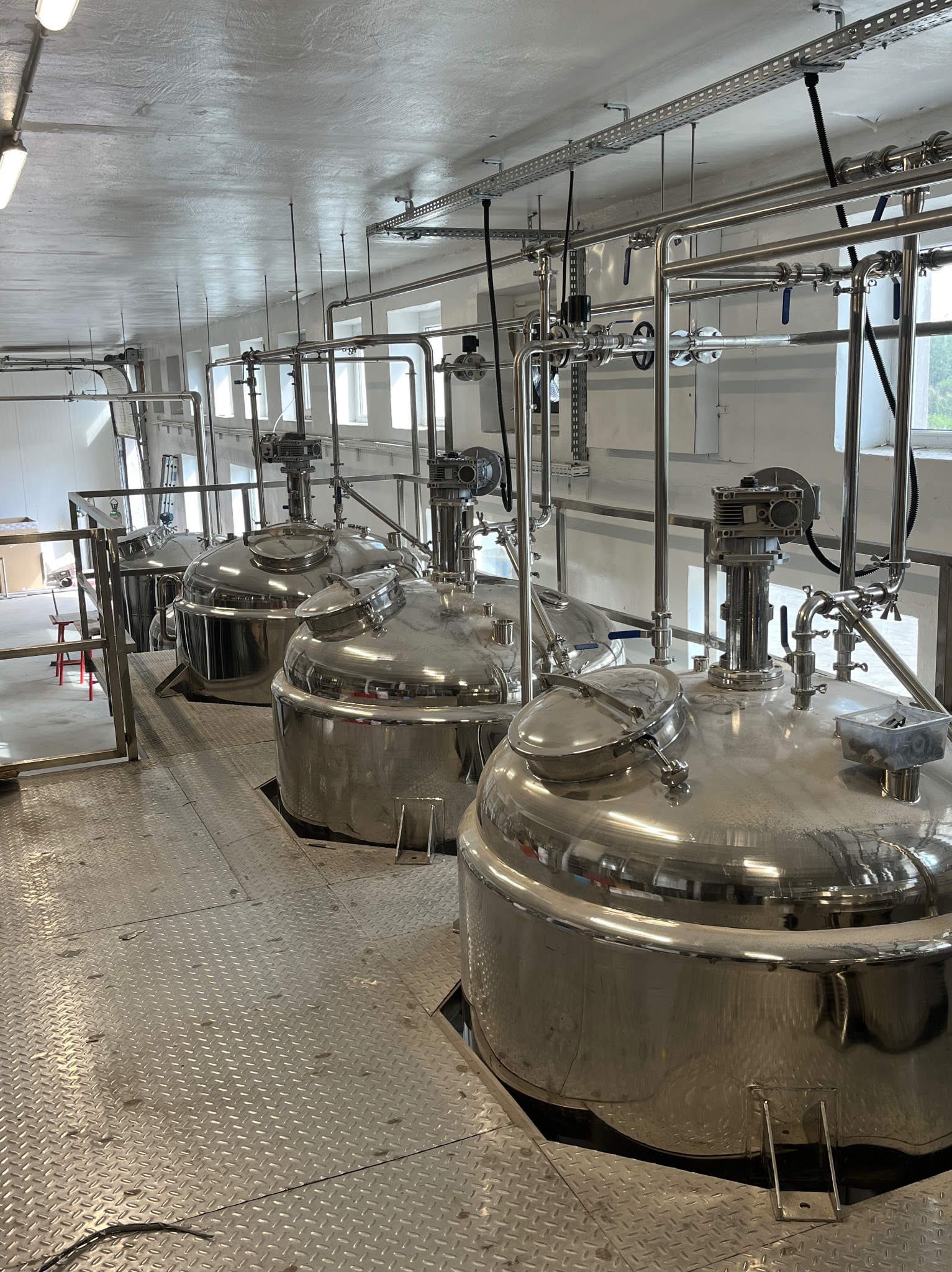 Wnętrze hali z czterema dużymi, metalowymi zbiornikami na platformie z blachy ryflowanej. Rurociągi i okna w tle. Widoczne elementy konstrukcji stalowej.