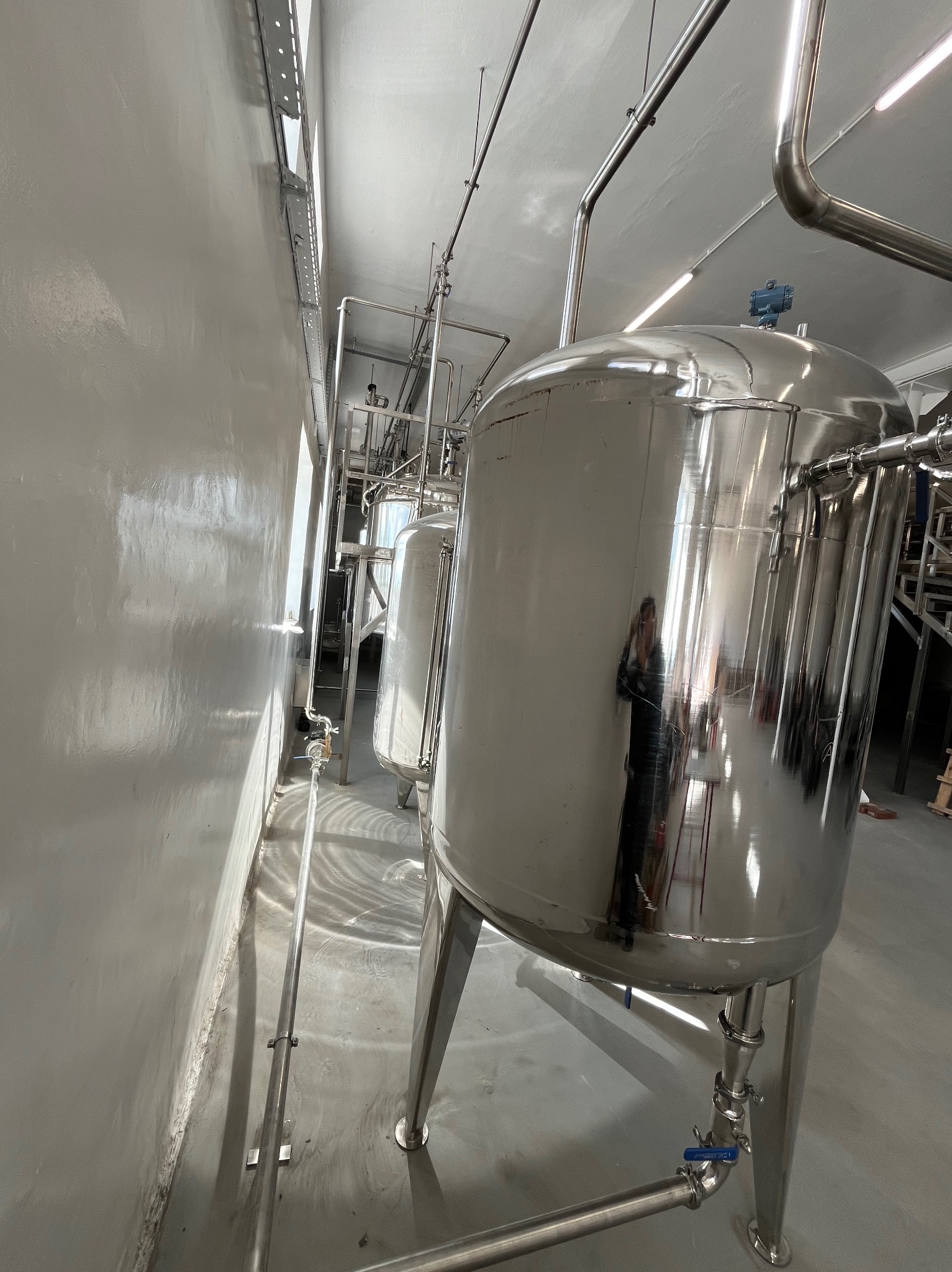 Dwa zbiorniki ze stali nierdzewnej w hali produkcyjnej w Wyszynie. Widoczne rury i armatura. Zbiorniki na trzech nogach, z połyskiem.