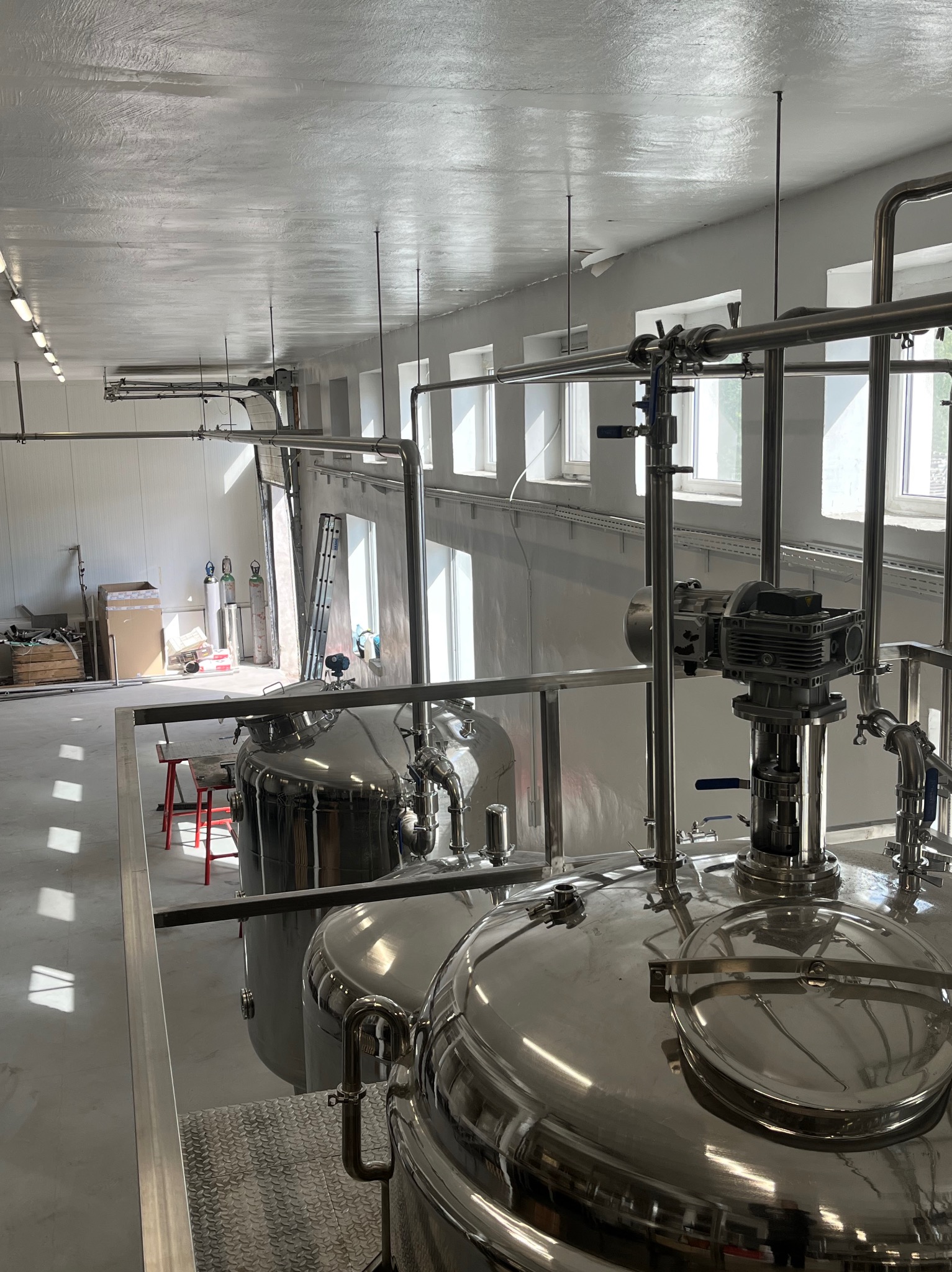 Wnętrze hali z dużymi, metalowymi zbiornikami i instalacją rur. Widoczne okna i światło dzienne. Elementy konstrukcyjne i wyposażenie przemysłowe w jasnej hali.