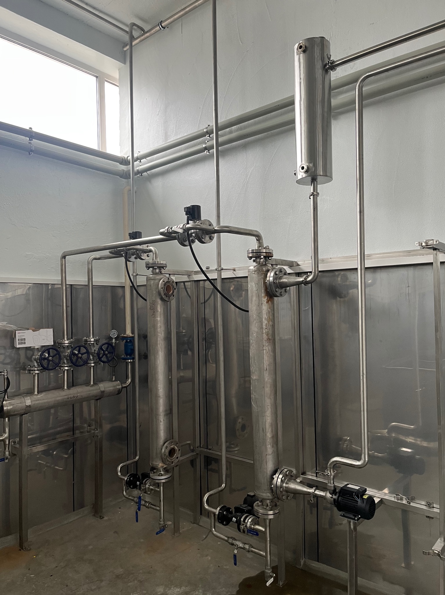 Instalacja przemysłowa z rurami ze stali nierdzewnej, zaworami i zbiornikami w pomieszczeniu z oknem. Widoczne złącza kołnierzowe i elementy mocujące.