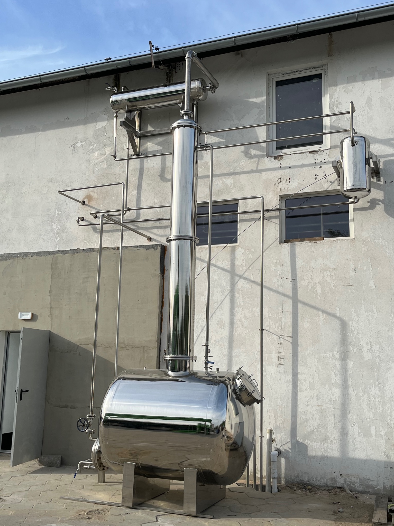 Instalacja przemysłowa ze stali nierdzewnej, zbiornik ciśnieniowy z rurami na tle budynku z oknami. Błyszcząca powierzchnia metalu odbija otoczenie.