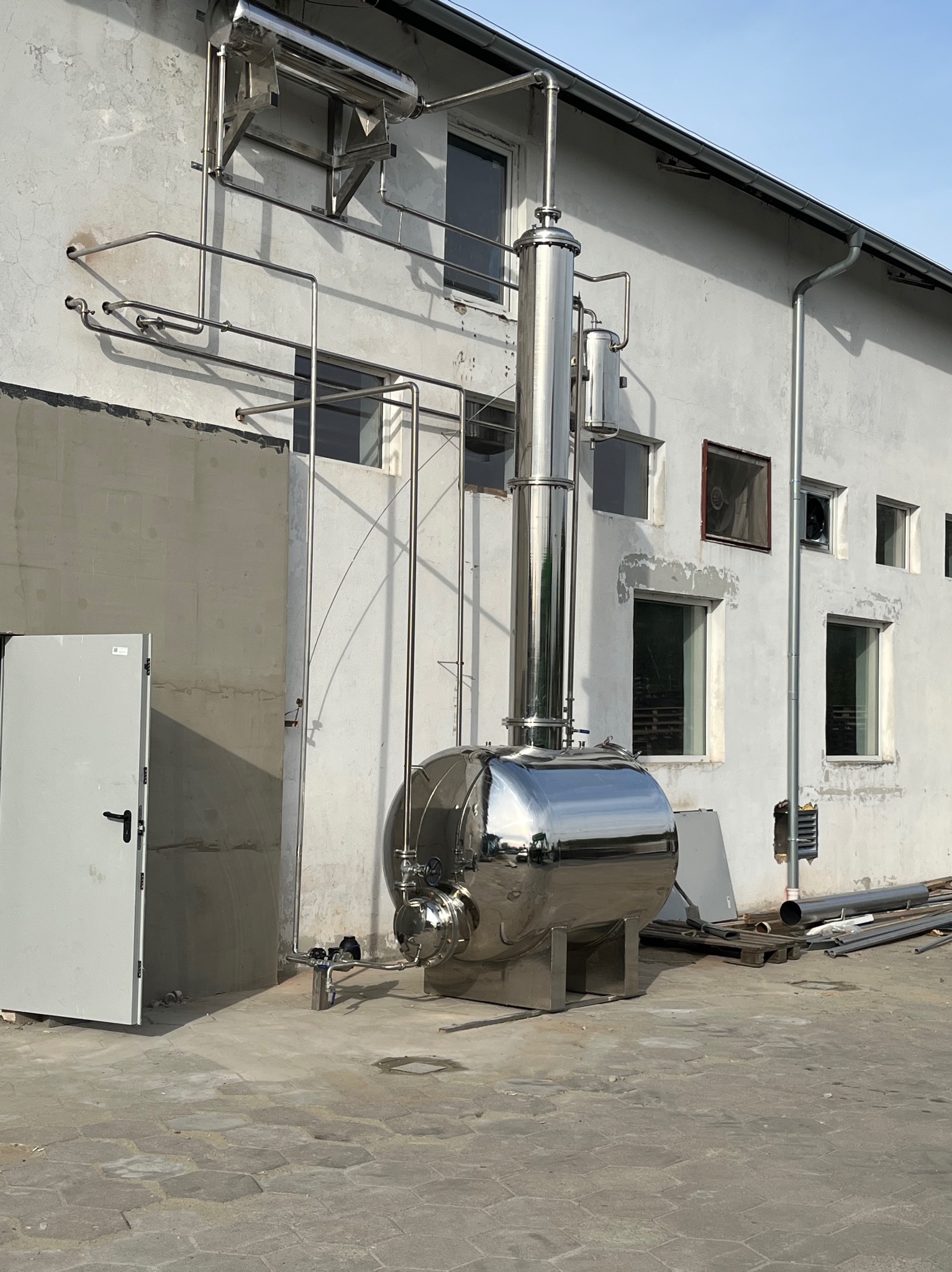 Błyszcząca, metalowa konstrukcja przemysłowa z rurami przymocowanymi do ściany budynku. Elementy konstrukcji spawane, widoczne okna i drzwi.