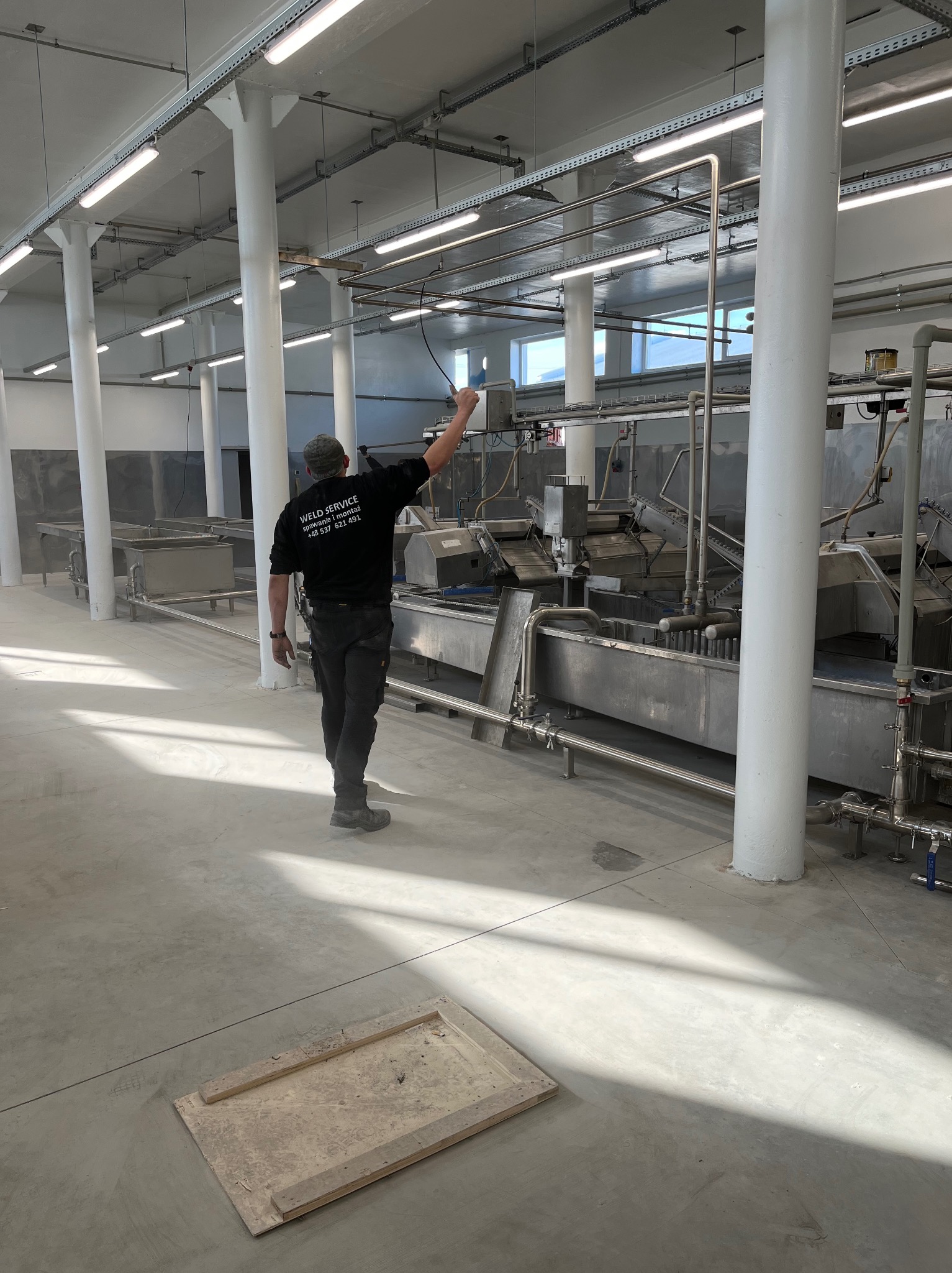 Pracownik w firmowej odzieży, kontroluje metalową instalację przemysłową w przestronnej hali z białymi kolumnami i oświetleniem. Widoczny fragment podłogi.