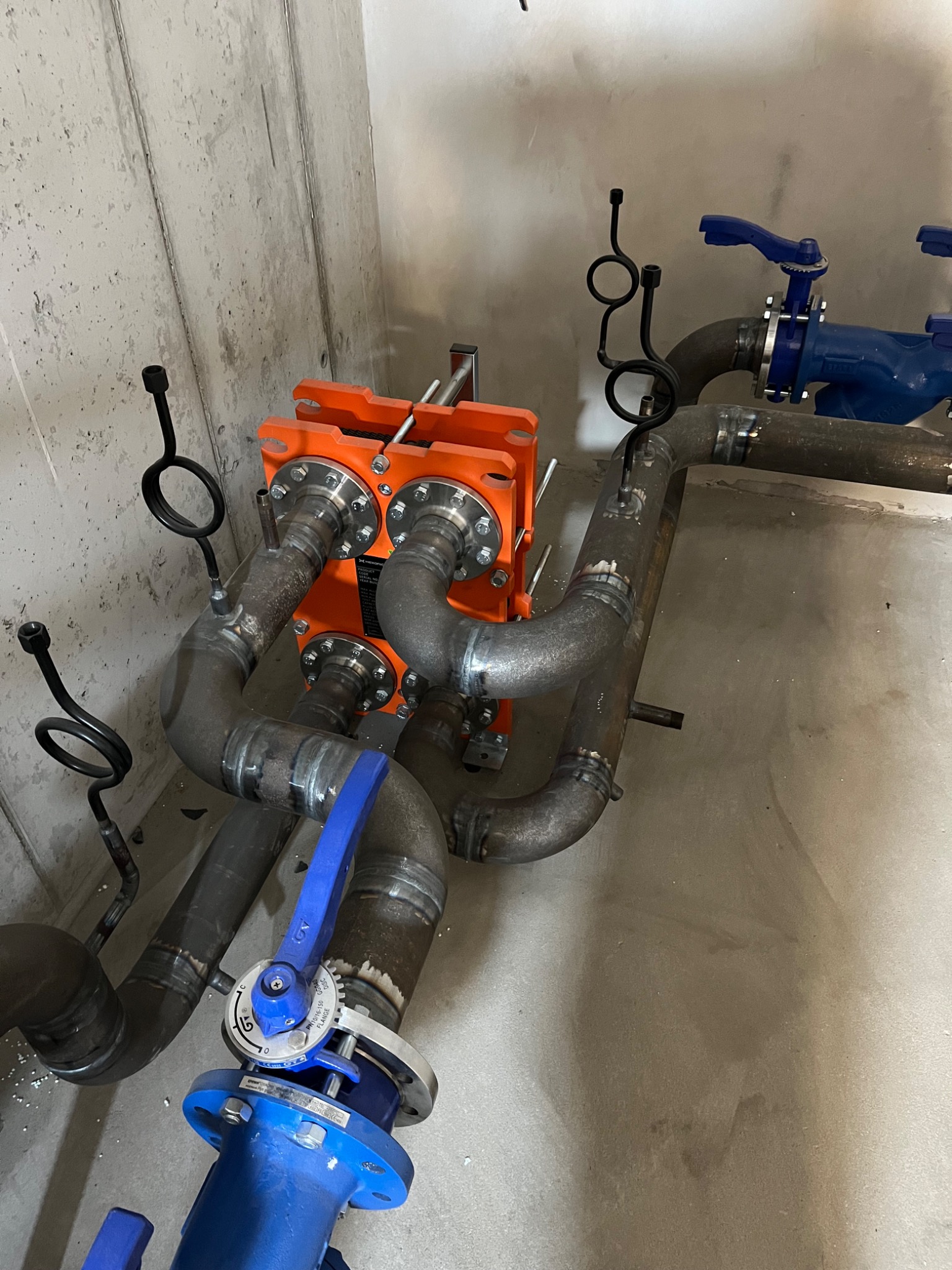 Pomarańczowy wymiennik ciepła i rury połączone spawami w kotłowni w Wyszynie, z widocznymi zaworami i elementami mocującymi.