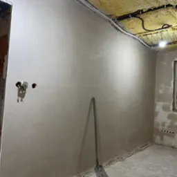 Świeżo otynkowane wnętrze z widocznymi przewodami elektrycznymi przy suficie i łopatą opartą o ścianę. Żarówka oświetla sufit z izolacją.