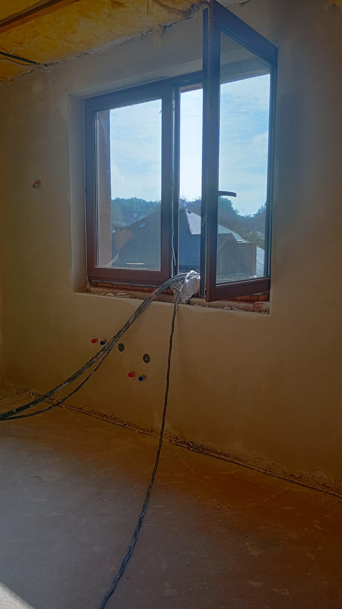 Wnętrze w trakcie prac wykończeniowych: ściany po nałożeniu tynku, otwarte okno z widokiem na zewnątrz, wystające kable instalacji. Widoczne ślady po tynkowaniu.