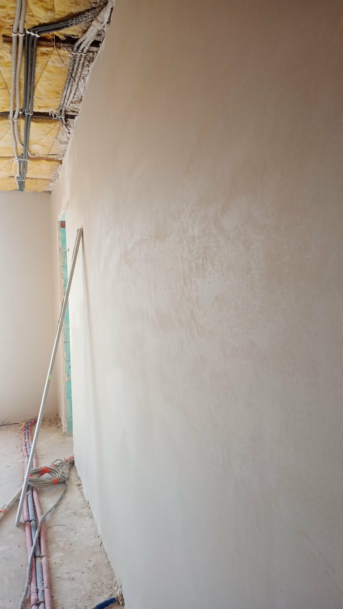 Świeżo nałożony, gładki tynk maszynowy na ścianie w trakcie remontu. Widoczne instalacje elektryczne i rurarskie oraz narzędzie do rozprowadzania tynku.