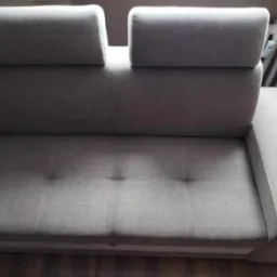 Szara, rozłożona kanapa z regulowanymi zagłówkami, widoczna z tyłu, z drewnianą podłogą i fragmentem grzejnika w tle.