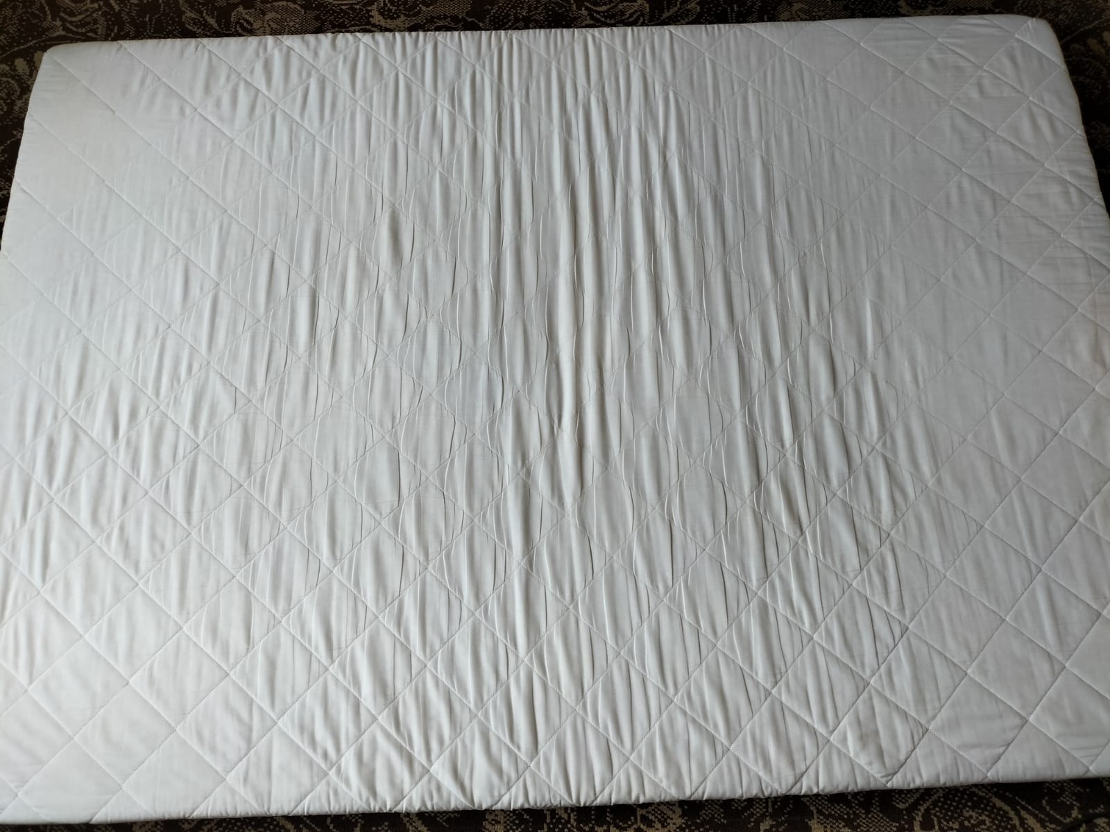 Biały, pikowany materac widziany z góry, leżący na ciemnym, wzorzystym dywanie.