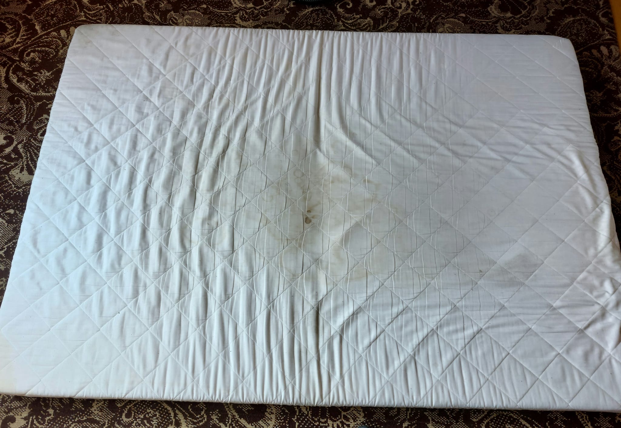 Biały materac z widocznymi plamami i zabrudzeniami leżący na wzorzystym dywanie, ukazujący stopień zużycia przed potencjalnym czyszczeniem.