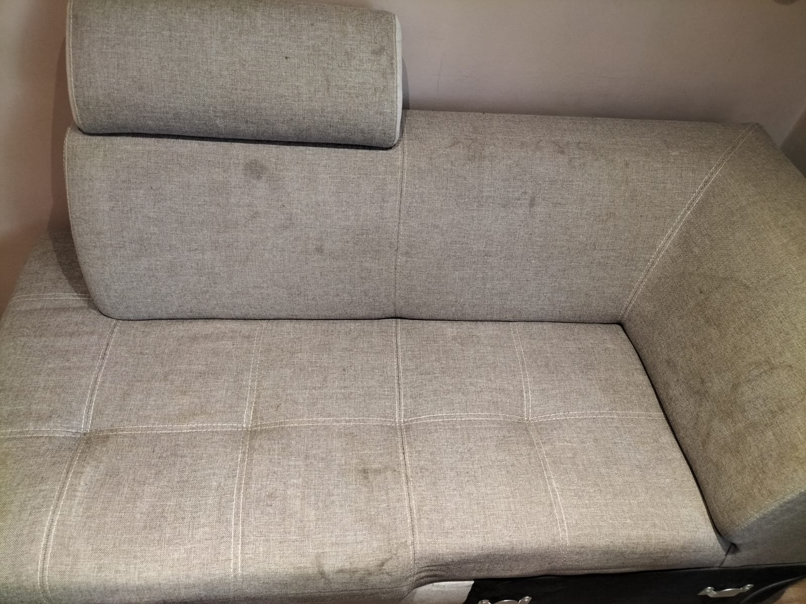 Szara, narożna kanapa z widocznymi plamami na siedzisku i oparciu, sfotografowana z góry w słabym oświetleniu.