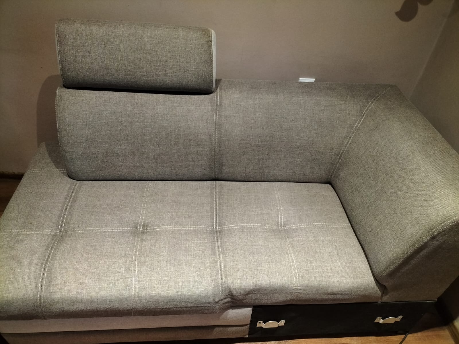 Szara, narożna kanapa z regulowanym zagłówkiem i pikowanym siedziskiem, widoczna szuflada na pościel z chromowanymi uchwytami.