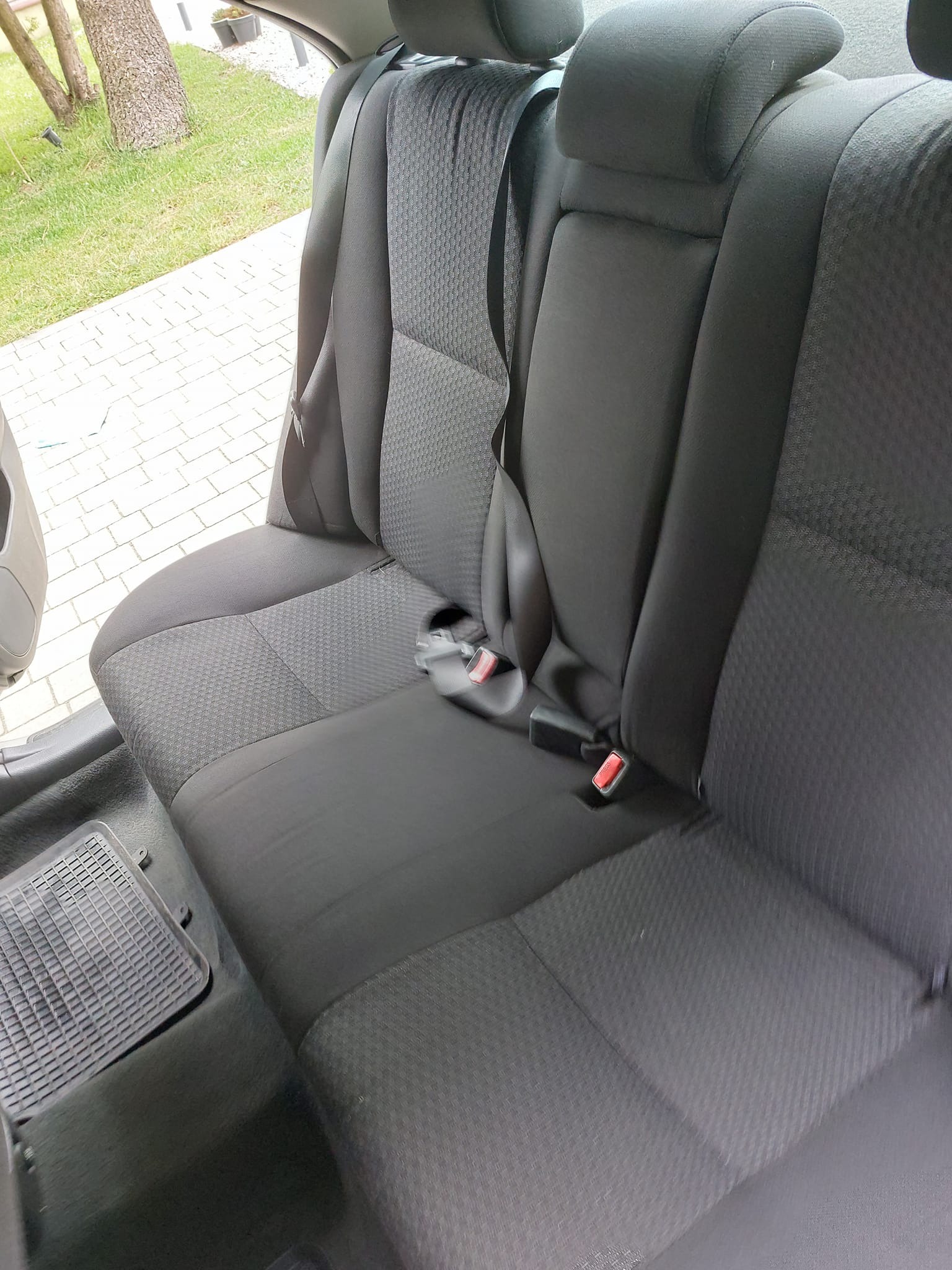Wnętrze samochodu: tylna kanapa z szarą tapicerką o drobnej fakturze, widoczne pasy bezpieczeństwa i gumowa wycieraczka na podłodze.