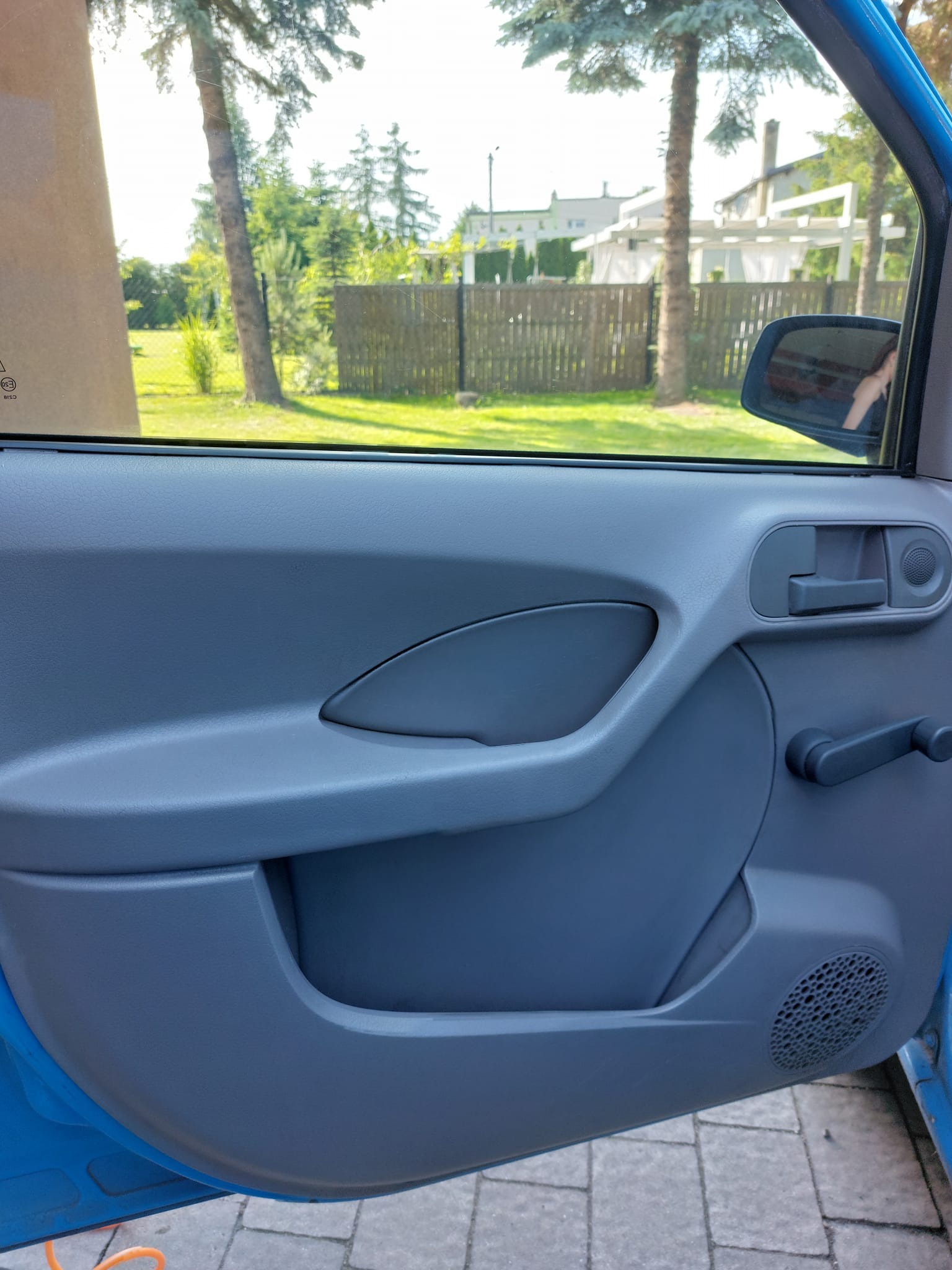 Wnętrze błękitnego samochodu, widok na szary panel drzwi z korbką do opuszczania szyby, na zewnątrz widoczne drzewa i budynki.