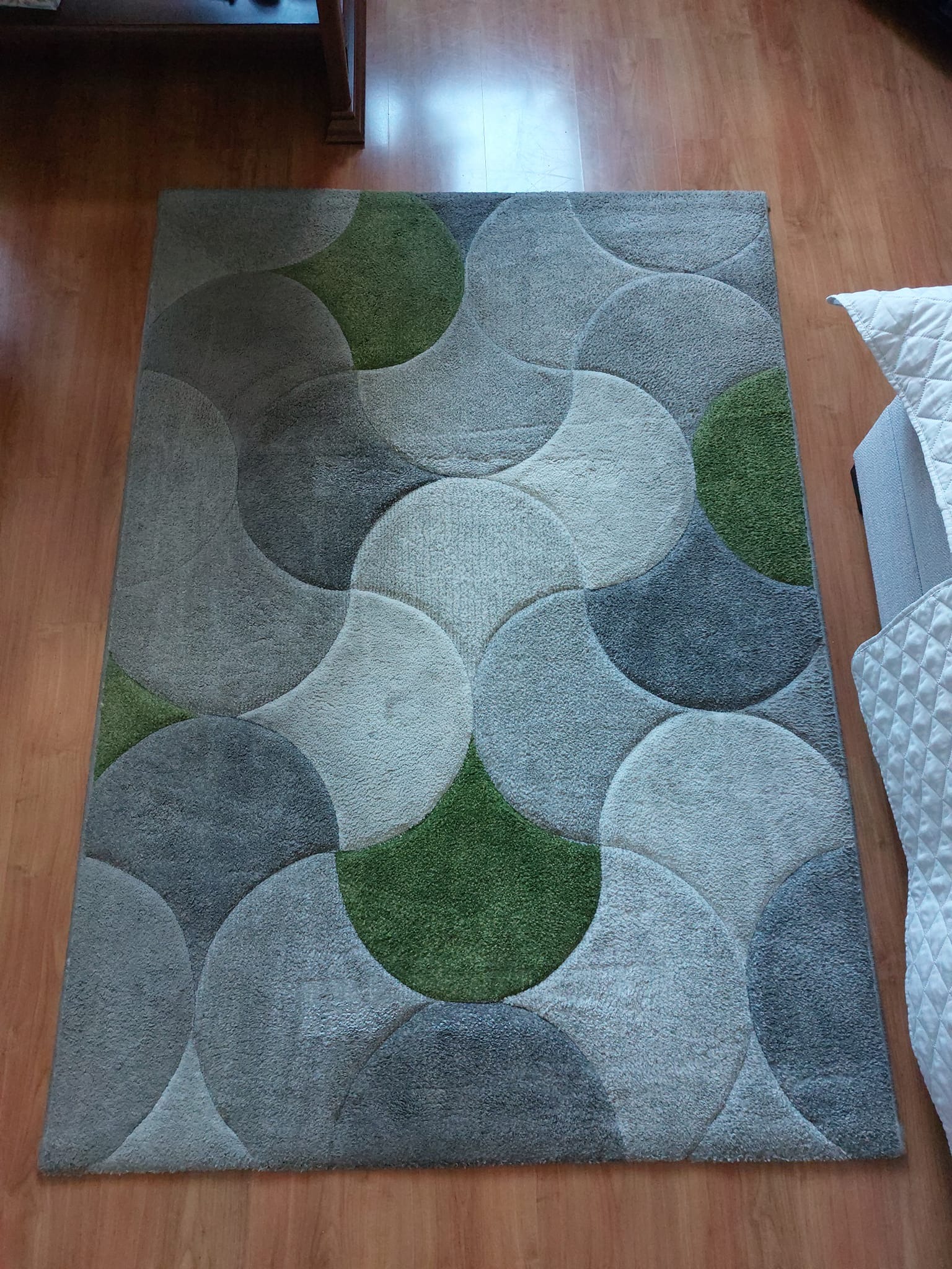 Prostokątny dywan z wzorem z nakładających się szarych i zielonych półokręgów leżący na drewnianej podłodze, fragment sofy z pikowaną narzutą widoczny w prawym górnym rogu.